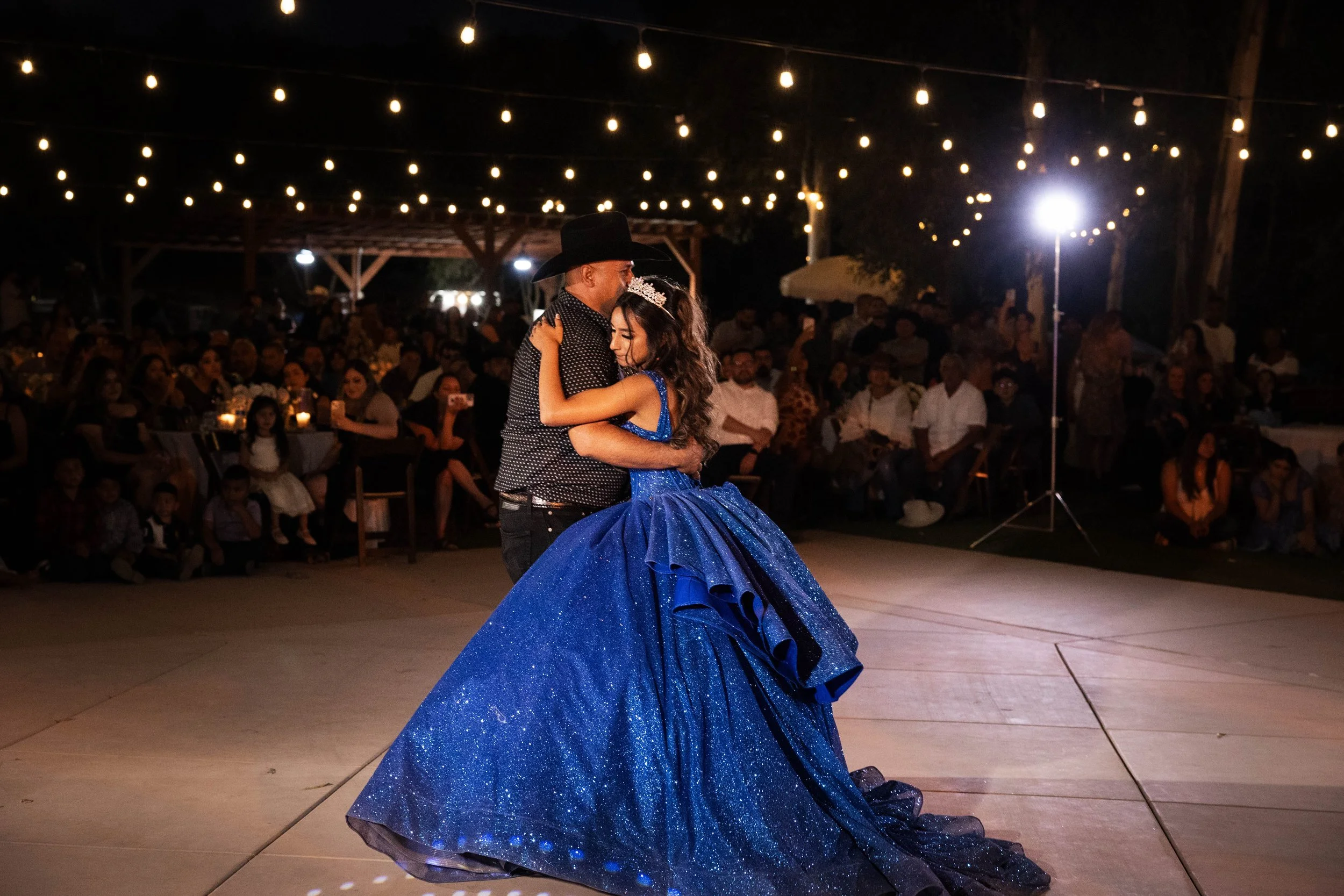 A young girl in a glittering blue gown and tiara dancing with a man in a black cowboy hat and dark shirt, at an outdoor event at night with string lights and a large audience watching.