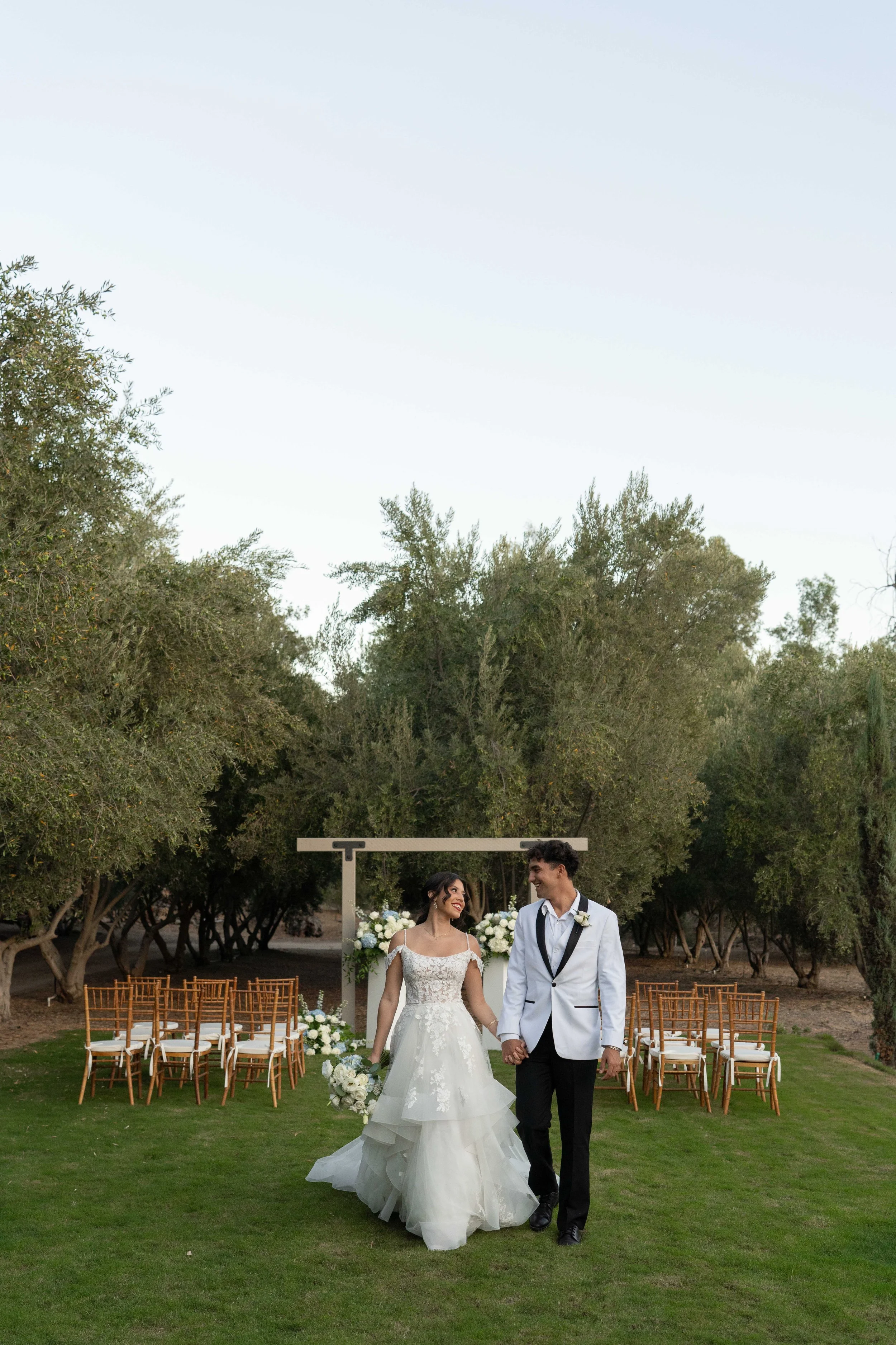 A bride and groom walk hand in hand on a grassy outdoor wedding venue, smiling at each other. The bride wears a white wedding dress and holds a bouquet, while the groom wears a white tuxedo jacket with black pants. Rows of wooden chairs are set up be