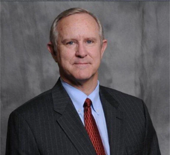 A middle-aged man in a dark pinstripe suit, light blue shirt, and red tie posing against a gray background.