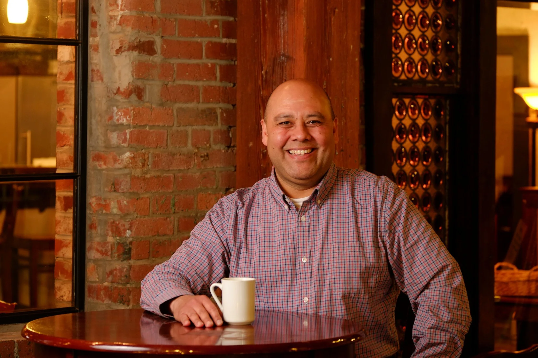 A smiling man with a bald head sitting at a round wooden table with a white mug in a cozy, warmly lit cafe with brick and wood interior features.