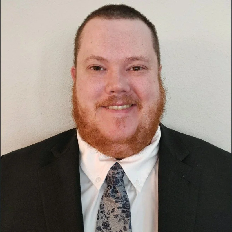 A man with a beard and short hair, dressed in a business suit and patterned tie, smiling at the camera against a plain background.