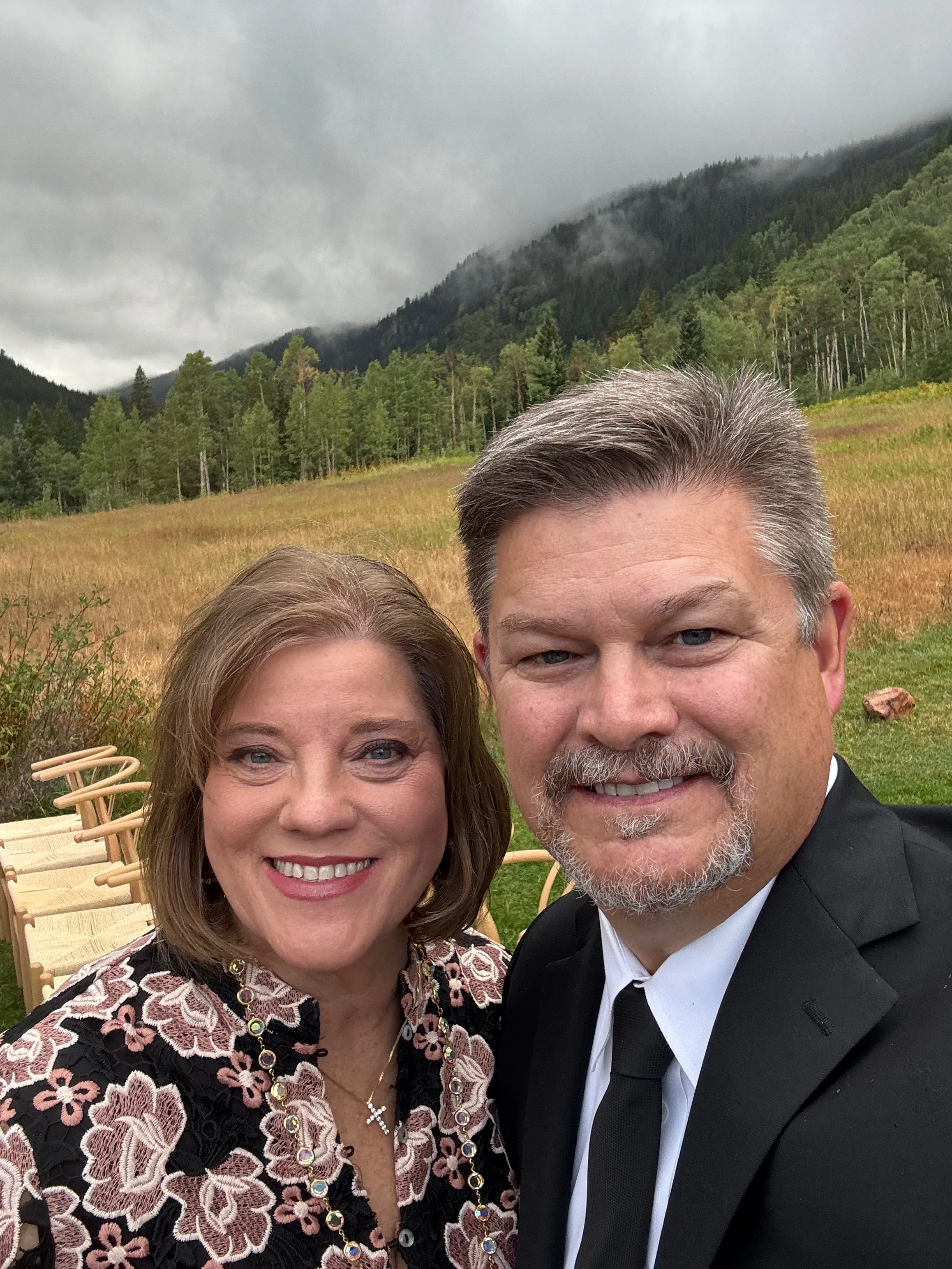 A smiling woman and man taking a selfie outdoors against a backdrop of a grassy field, trees, and mountain with clouds.