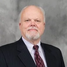 A professional headshot of an older man with a white beard and mustache, wearing a dark suit, white shirt, and patterned tie, against a gray background.