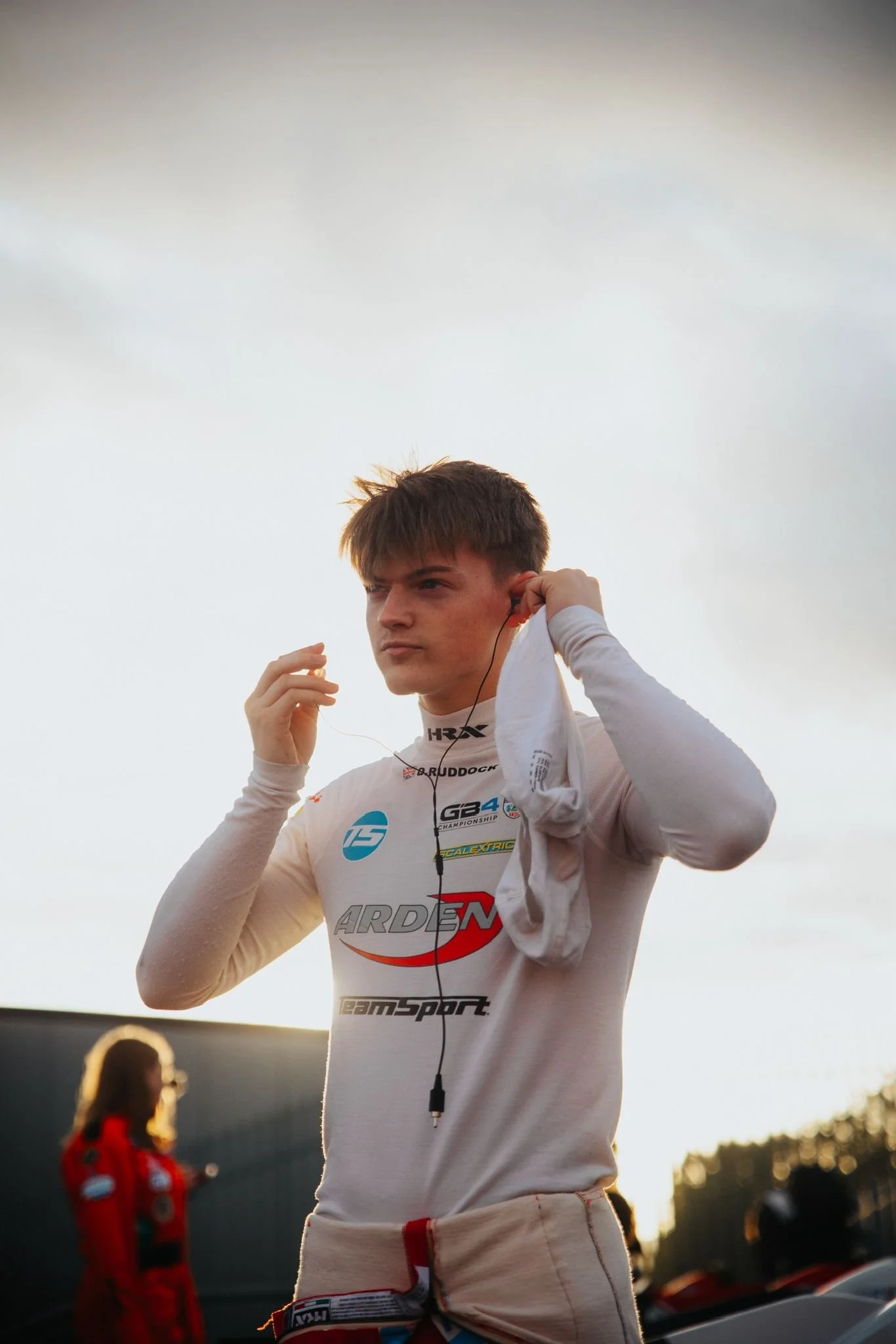 A young male race car driver adjusting his hearing protection outdoors during sunset, wearing a racing suit with logos.