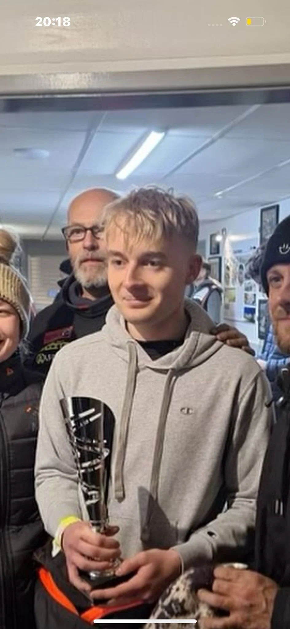 A young man in a gray hoodie holding a trophy, surrounded by smiling people in a social indoor setting.