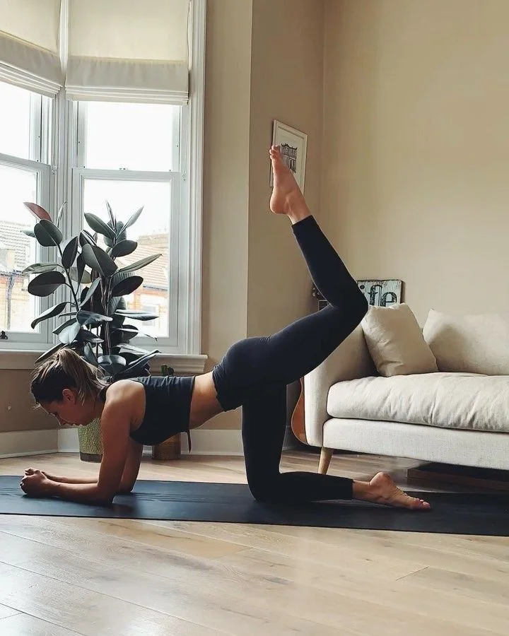 A woman is practicing pilates in her living room, elbows on mat, leg lifted.