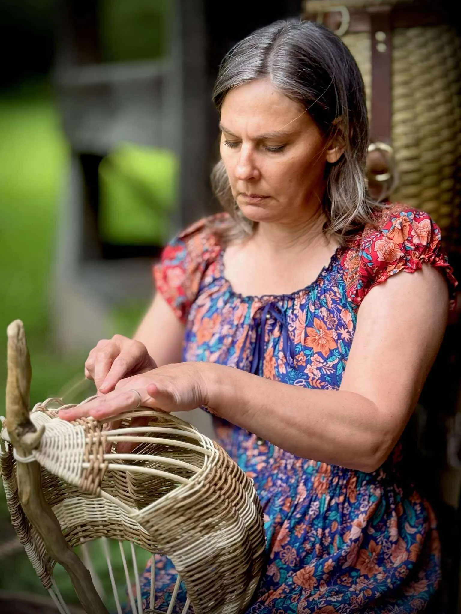 A woman with gray hair, wearing a colorful floral dress, working on a wicker basket outdoors.