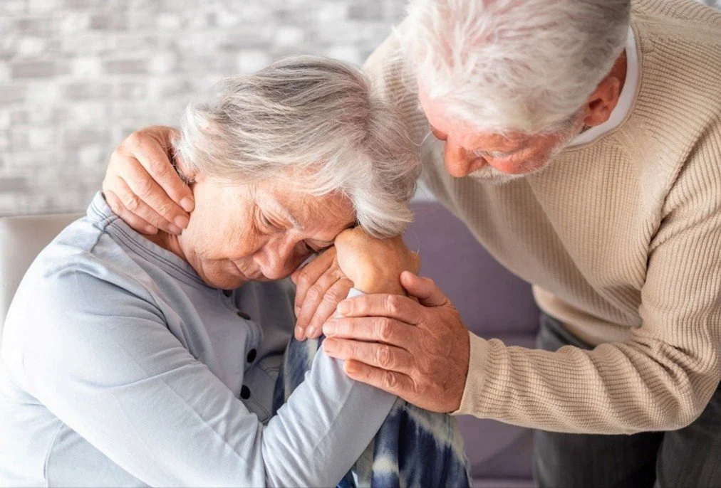 An elderly woman holds her hand and forehead against a man's face while he comforts her, showing an emotional moment of support and care.
