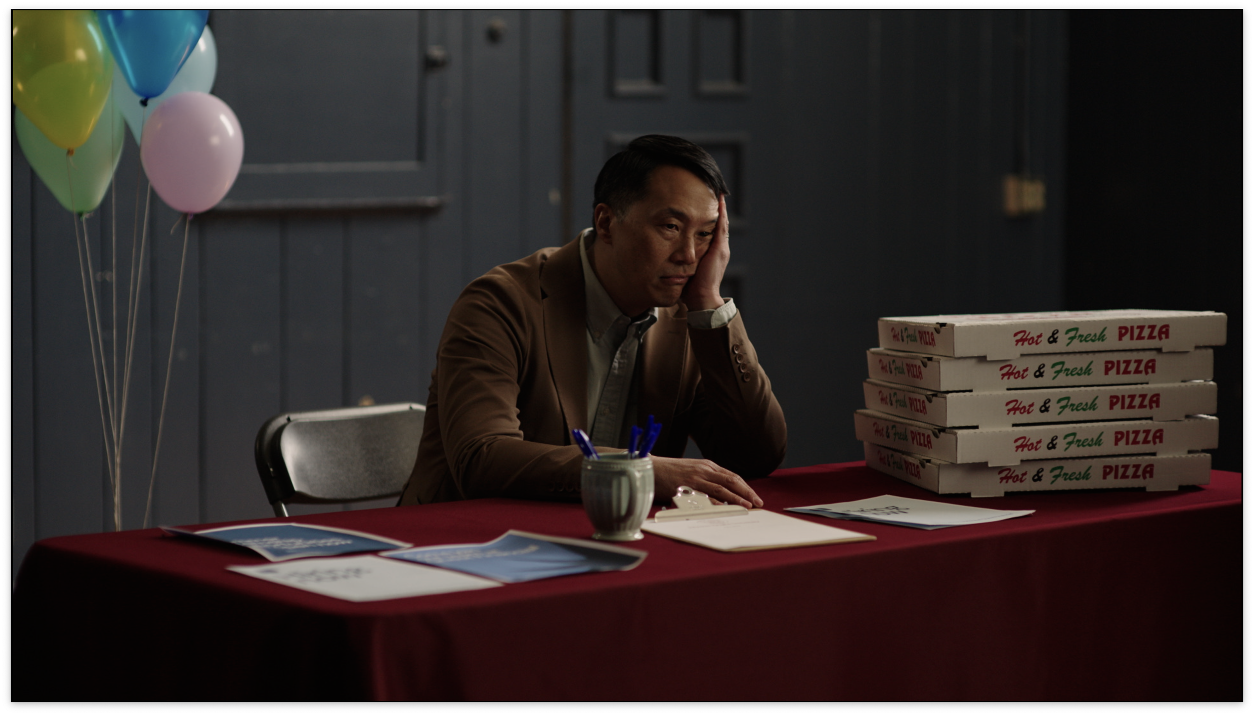 A man sitting at a table with six pizza boxes, looking disappointed or tired, resting his head on his hand. There are balloons, brochures, a mug with pens, and a clipboard on the table.