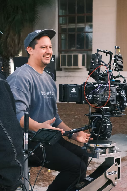 A man wearing a gray sweatshirt and black cap sits behind a professional film camera setup outdoors. He is smiling and appears to be in a conversation or interview, with a building and palm tree in the background.