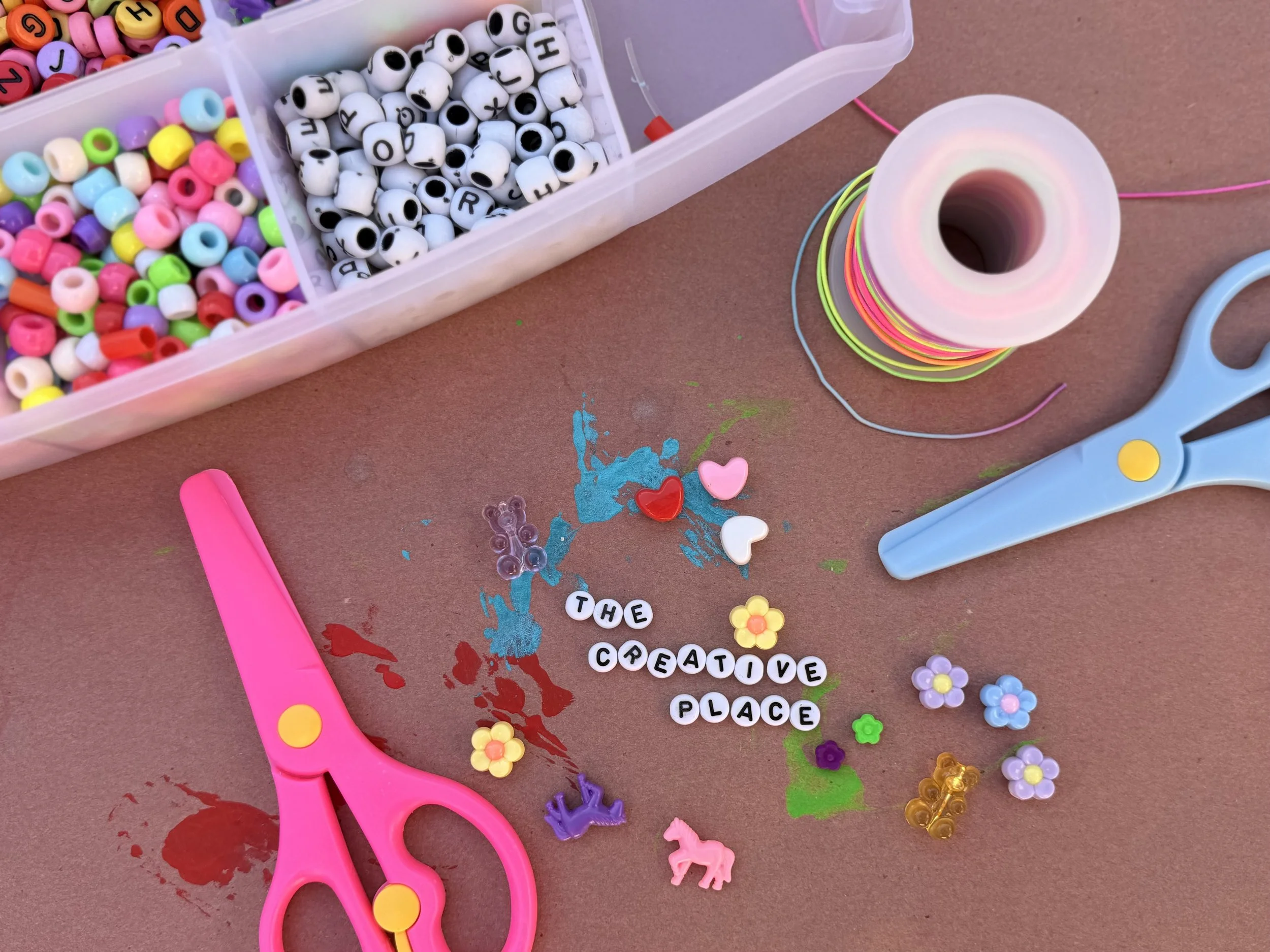 Craft supplies including colorful beads, alphabet beads, plastic scissors, embroidery thread, and decorative charms on a craft table.