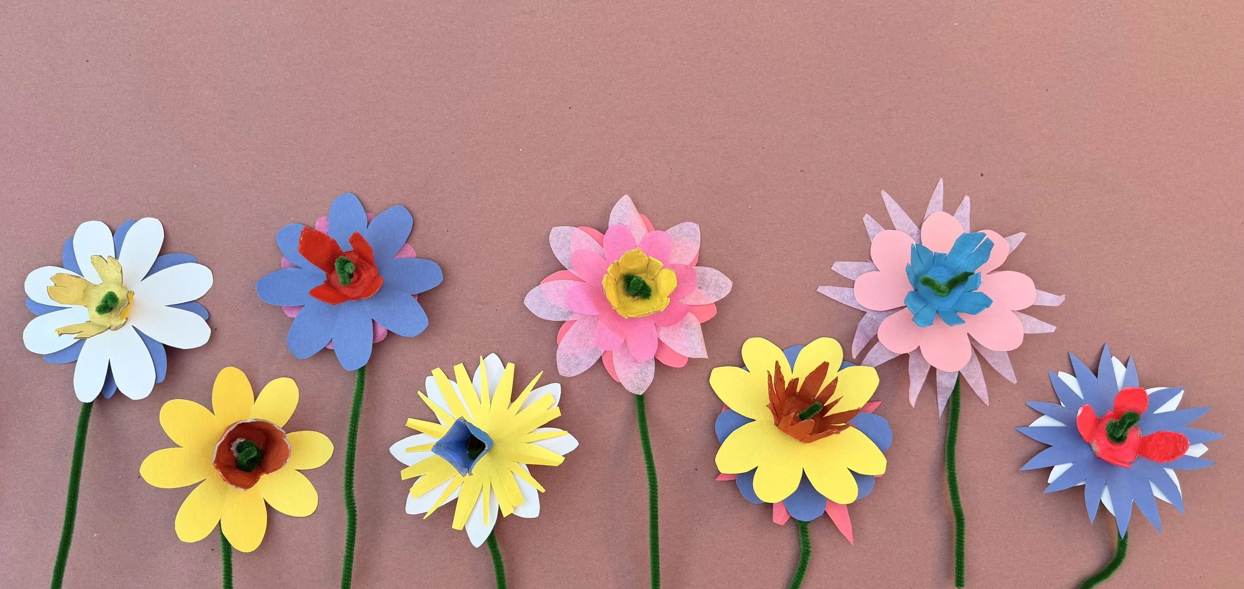 Colorful paper flowers with pipe cleaner stems arranged on a pink background.