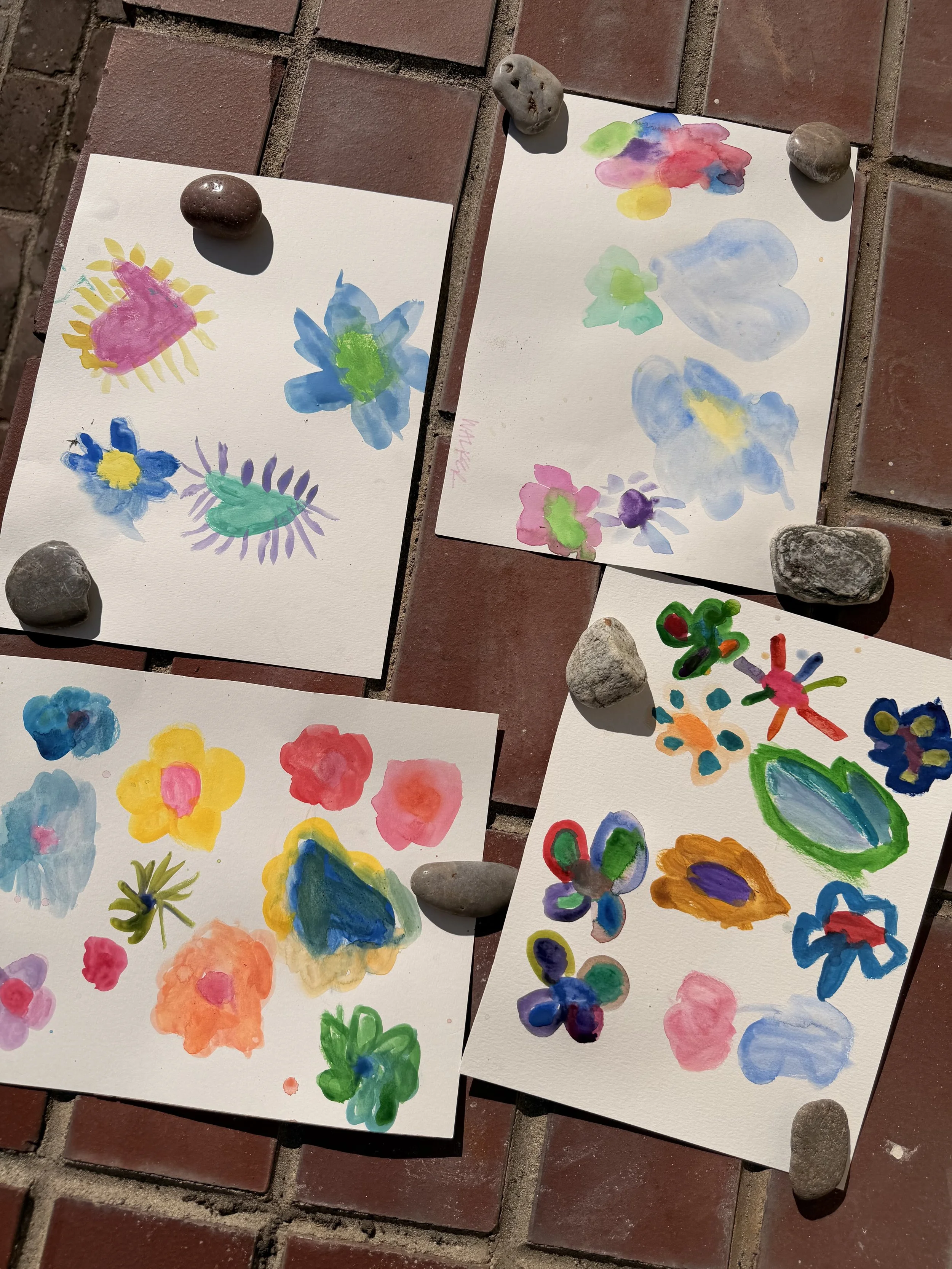Four watercolor paintings of flowers and clouds, displayed on a brick surface with small rocks holding down each painting.