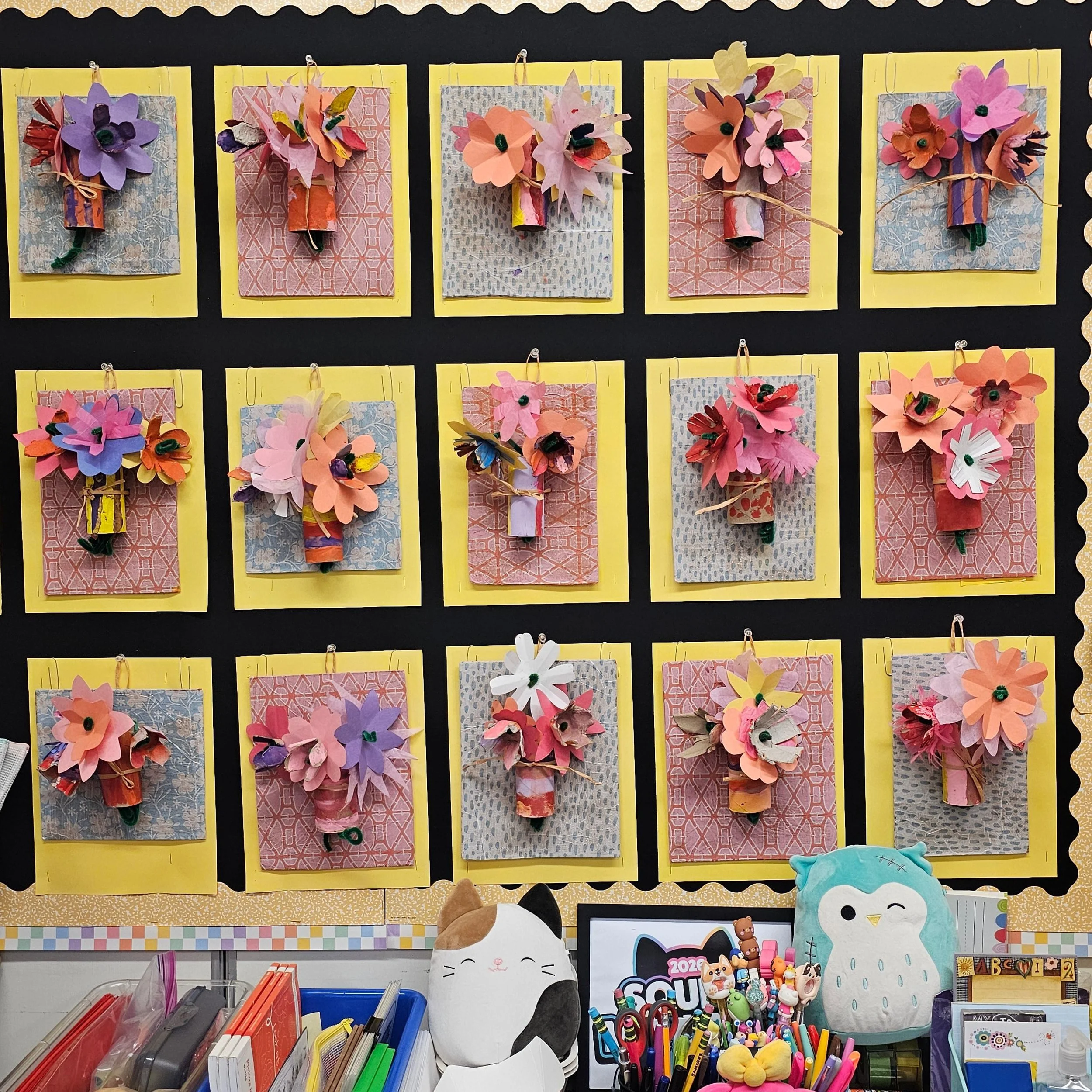 A bulletin board display with 15 paper flower crafts arranged in a grid, each mounted on colorful backgrounds with yellow borders, and decorated with various embellishments. Below the board are shelves filled with school supplies, plush toys, and other classroom items.
