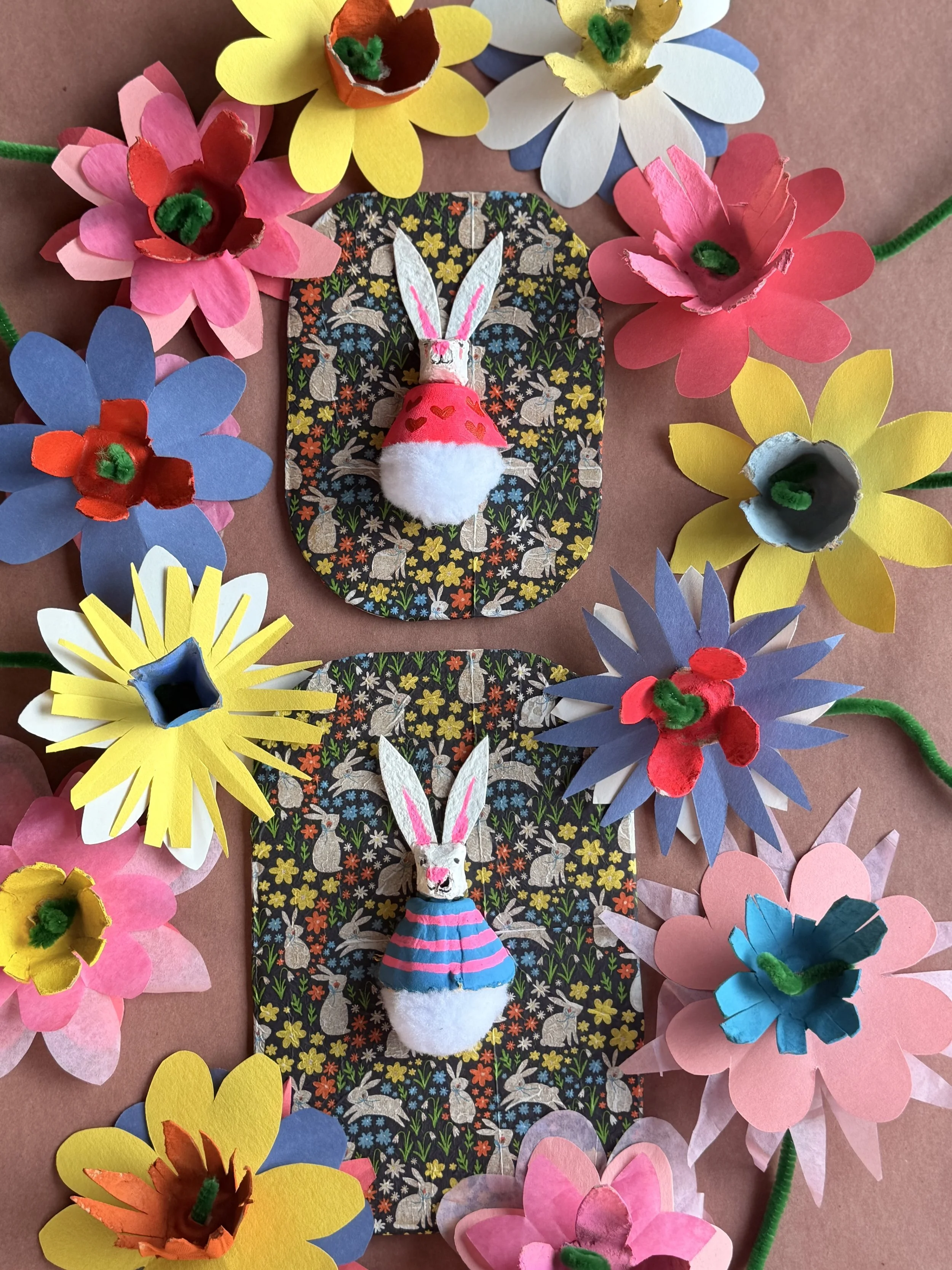 Craft display featuring two rabbit puppet characters on black patterned background and surrounded by colorful paper flowers with pipe cleaner stems.