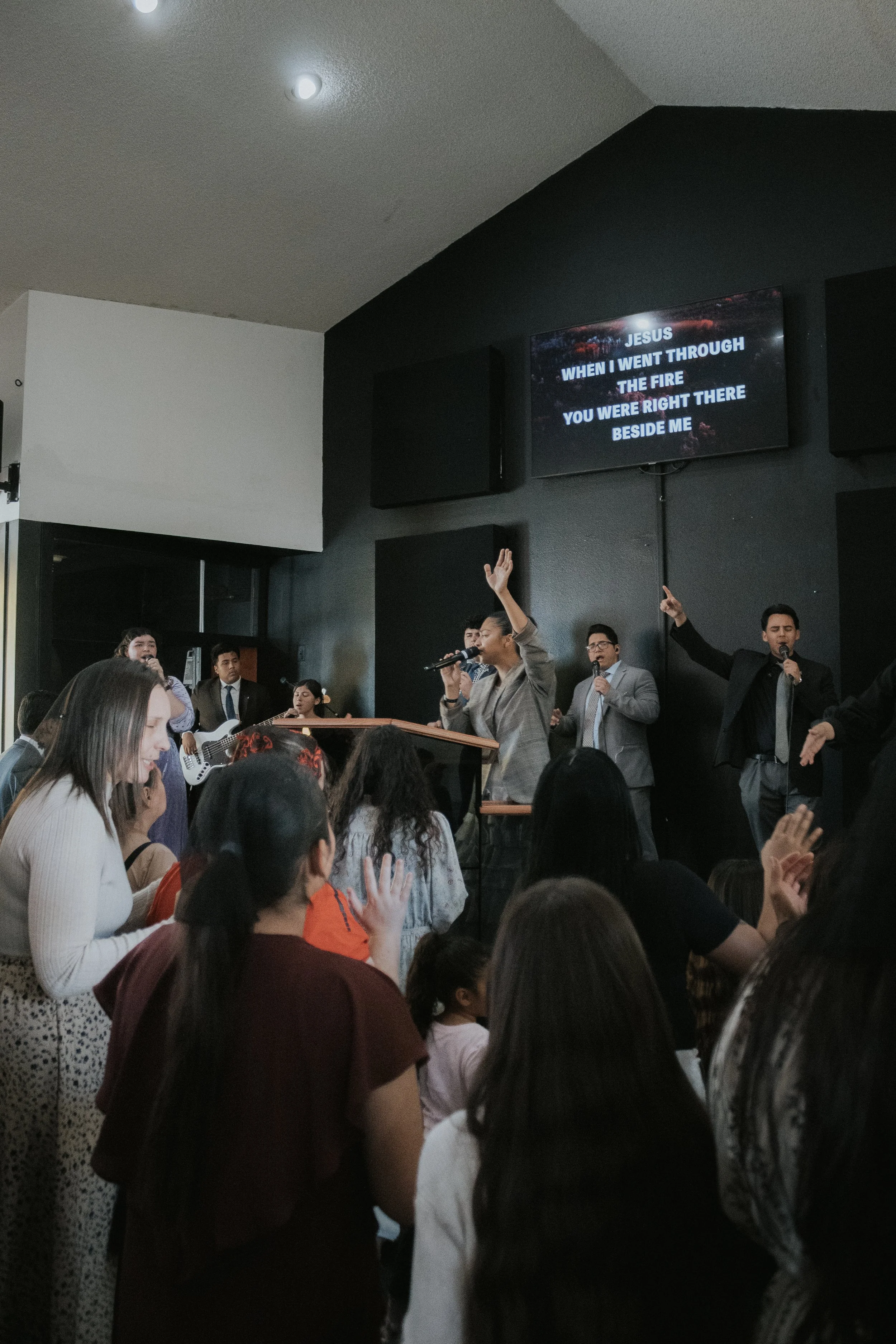 Group of people singing and praying during a church service, with a screen displaying lyrics behind them.