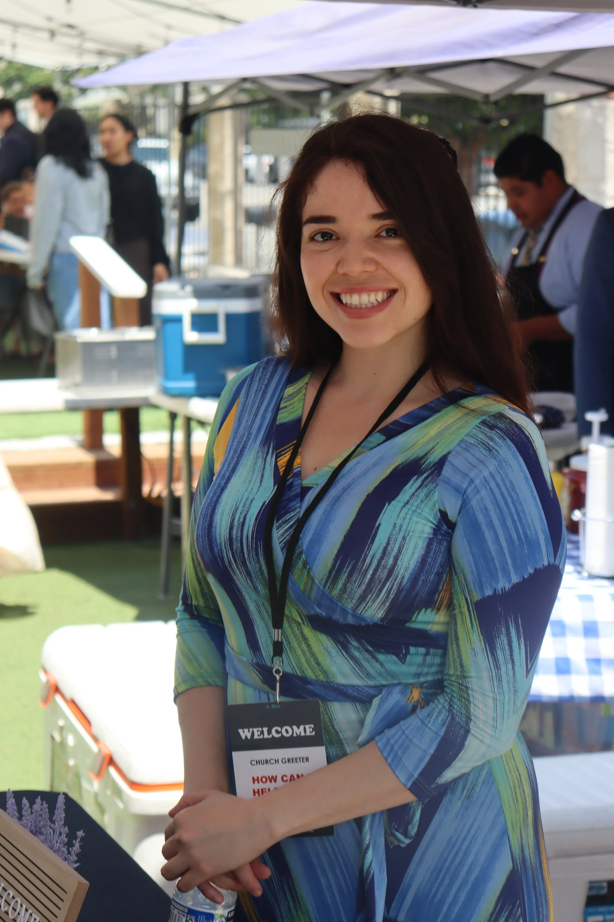 A smiling young woman with shoulder-length brown hair and fair skin wearing a colorful patterned dress and a church greeter badge at an outdoor event.