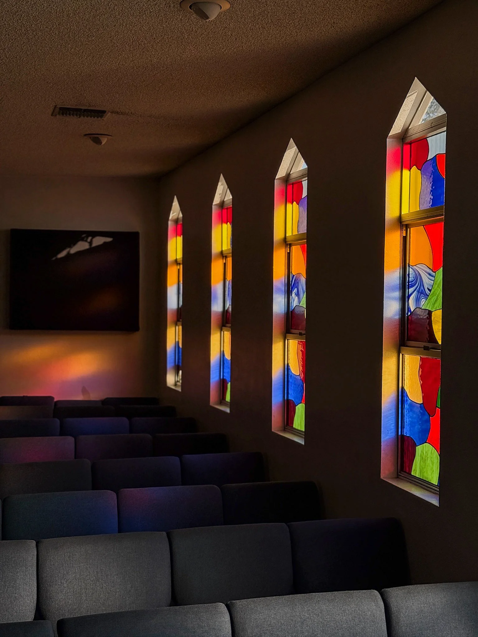 Sunlight passing through colorful stained glass windows in a quiet room with empty gray chairs and a dark wall-mounted television.