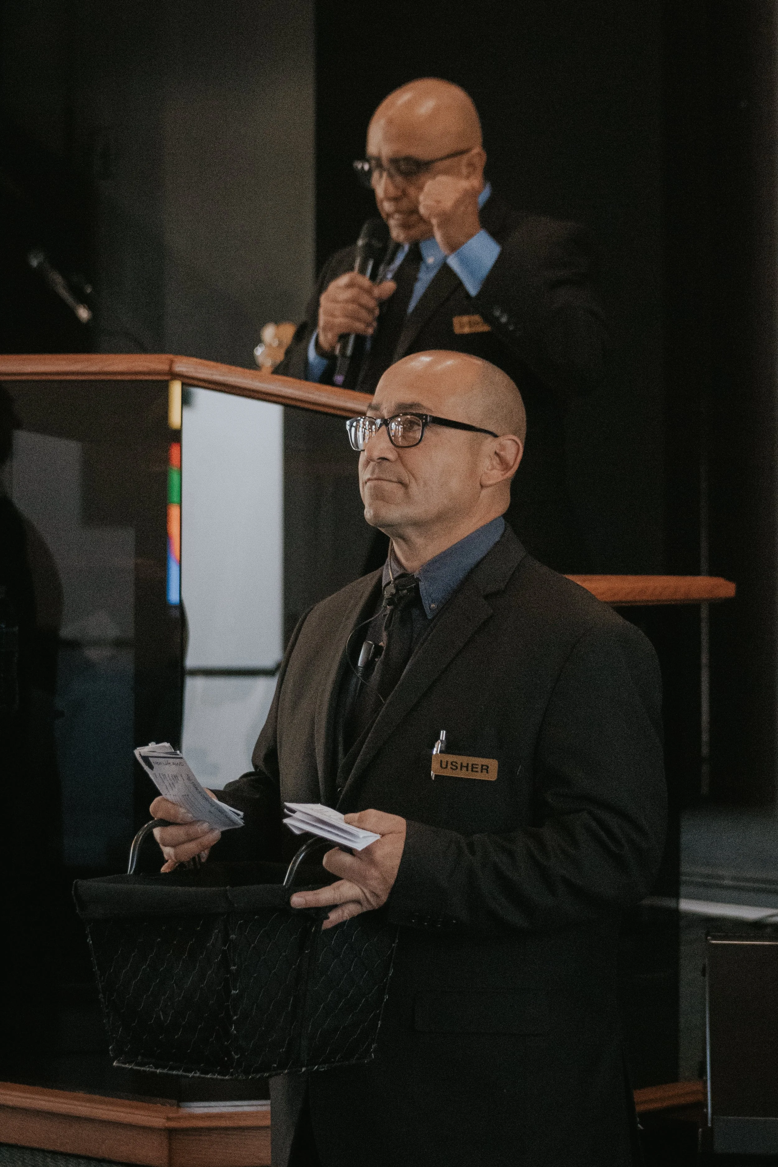 A man wearing glasses, a suit, and a name tag reading 'Usher' stands in front of a crowd holding a basket and papers, with a man speaking at a podium in the background.