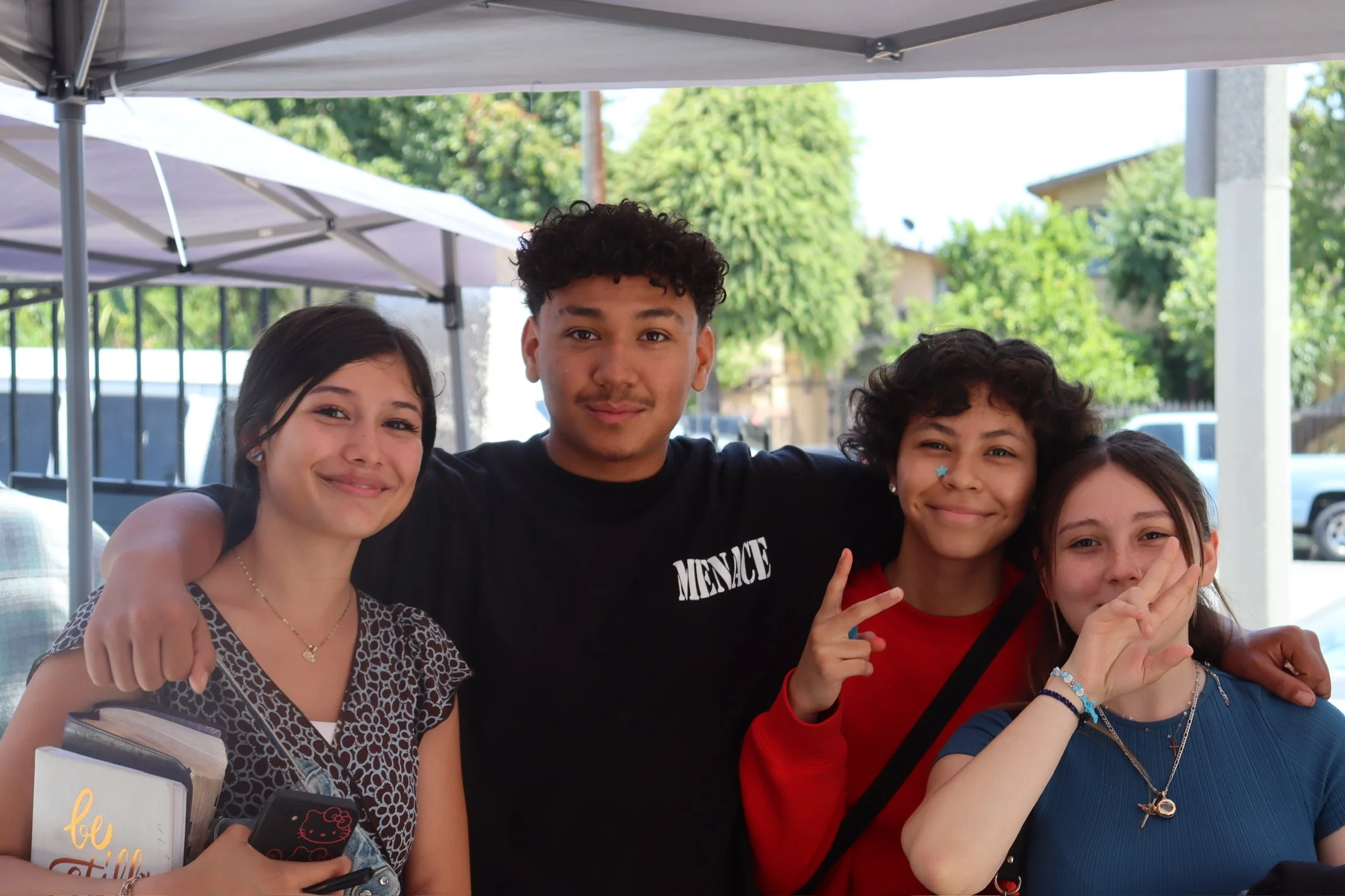 Four teenagers smiling and posing together outdoors, with tents and trees in the background.
