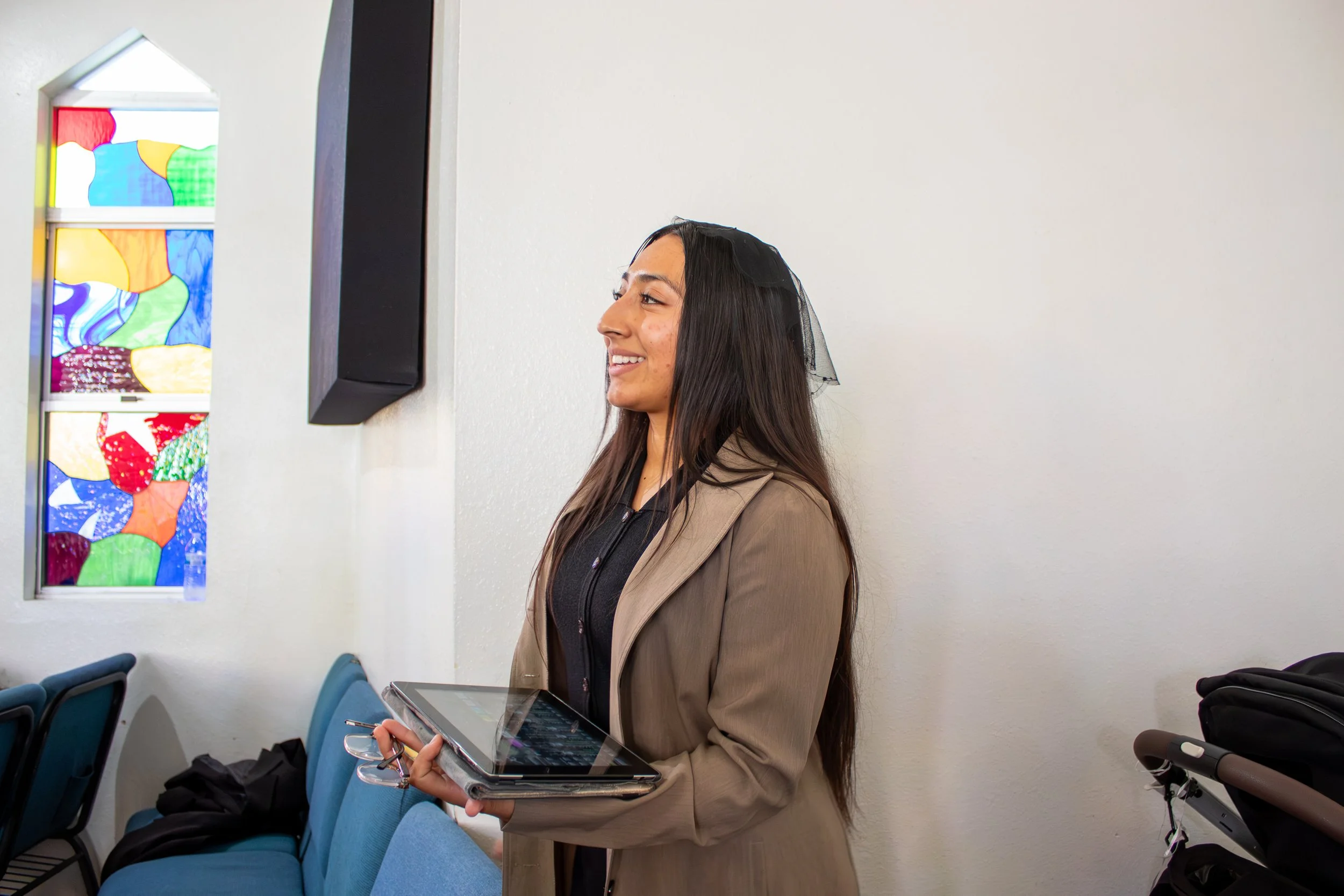 A woman with long dark hair and a tan jacket standing indoors, holding a tablet and glasses, smiling, in front of a white wall and colorful stained glass window.