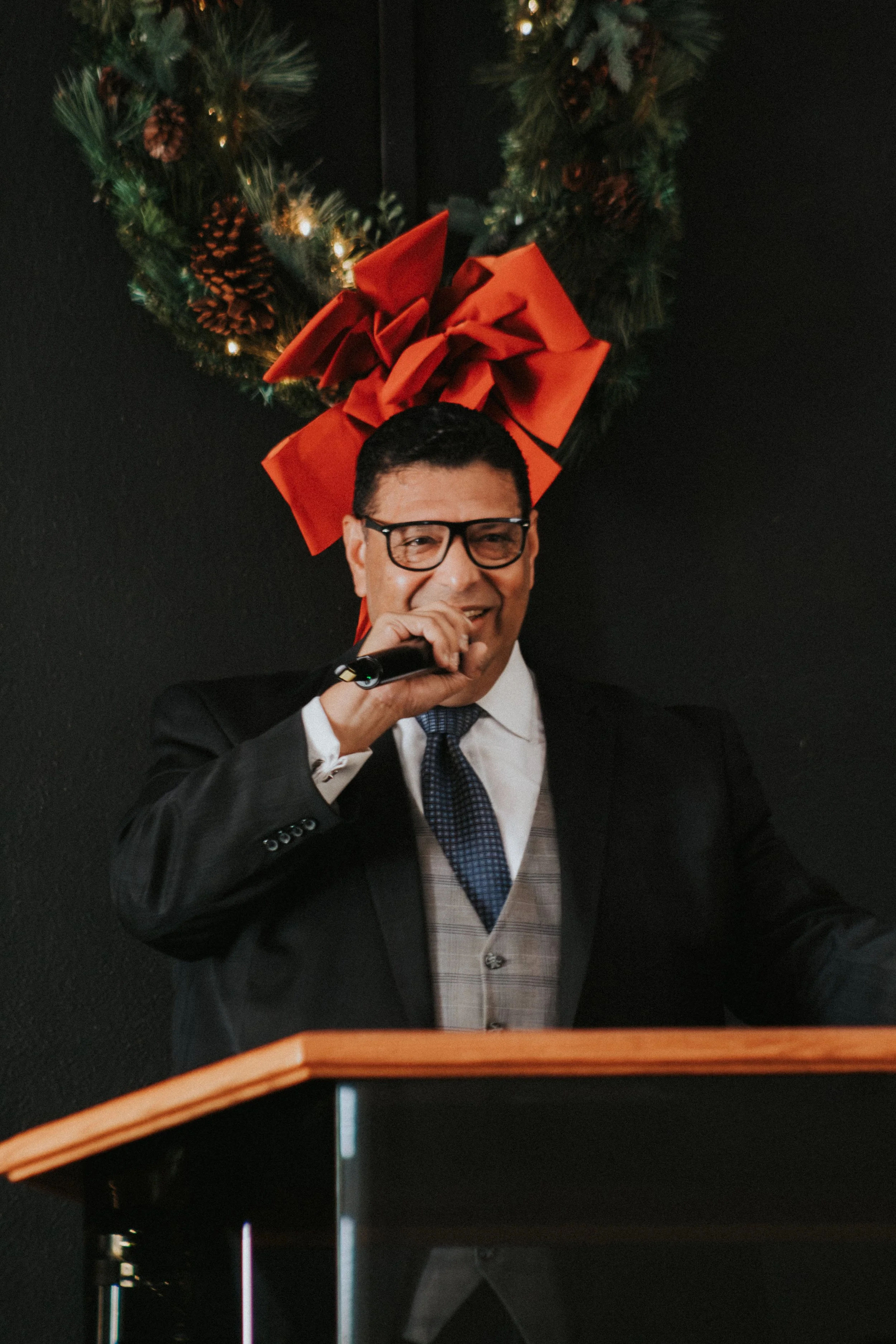 A man in a suit speaking at a podium during a holiday event, with a Christmas wreath and large red bow in the background.