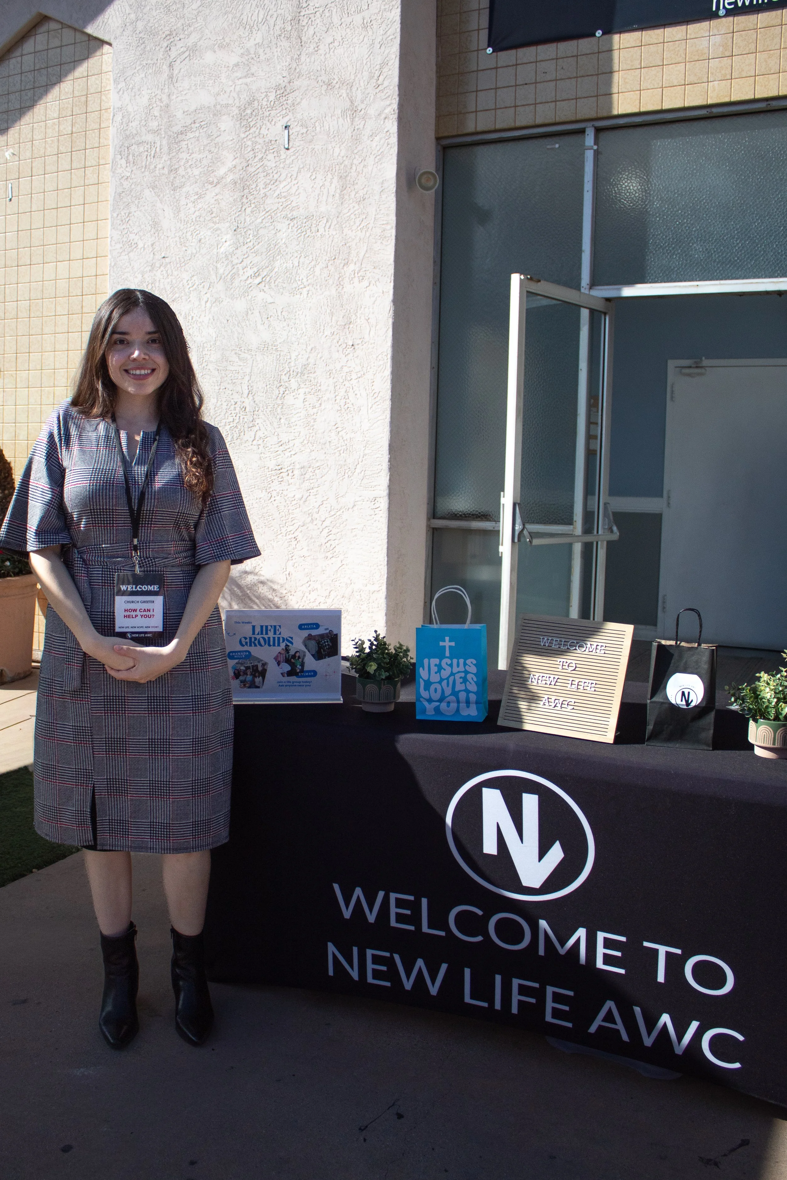 A young woman standing beside a black table with a welcome sign for New Life AWC, decorated with small plants, gift bags, and signs related to a church or community event outside a building.