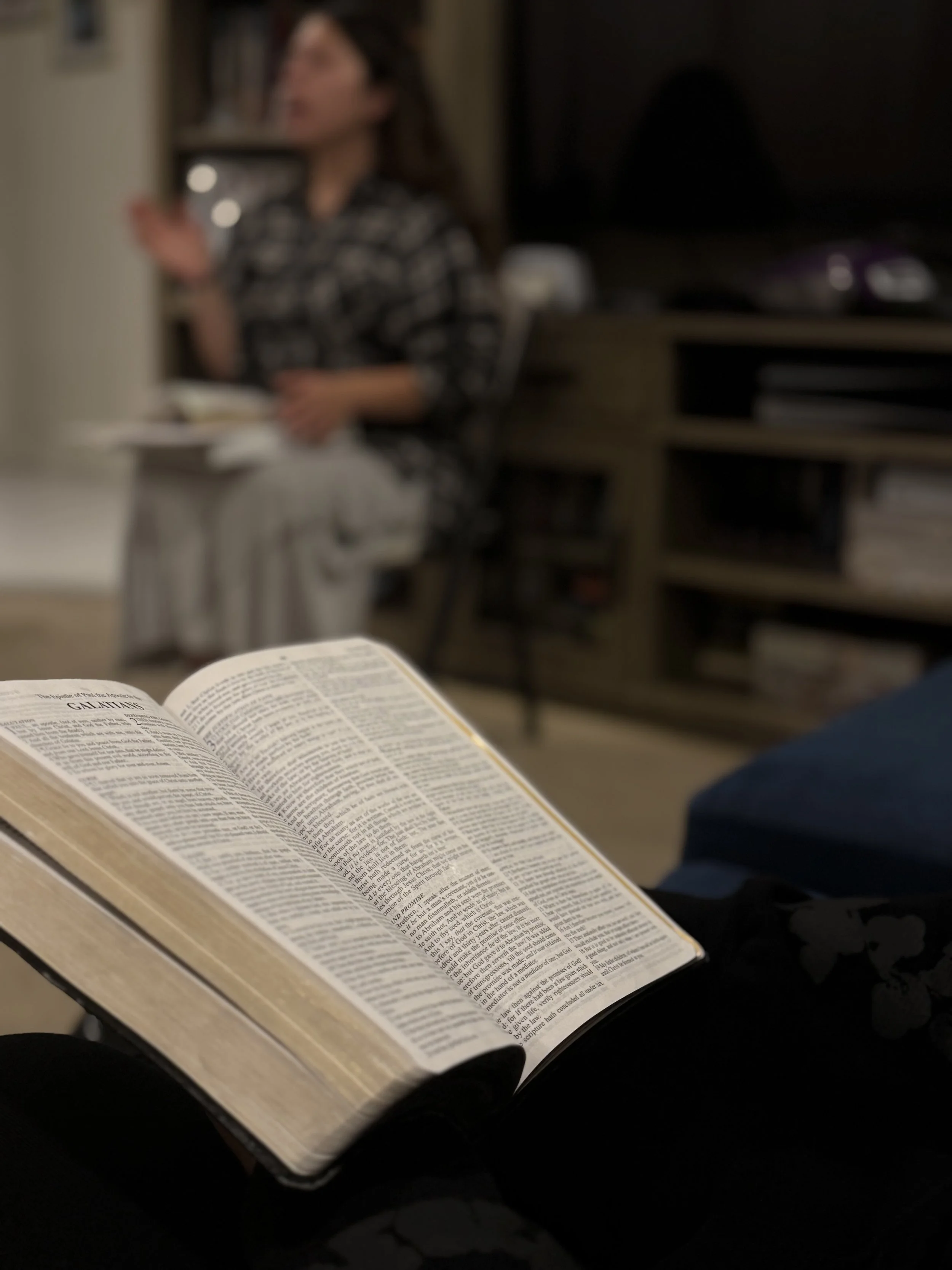 Open Bible resting on a person's lap in focus, with a woman sitting in a living room blurred in the background.