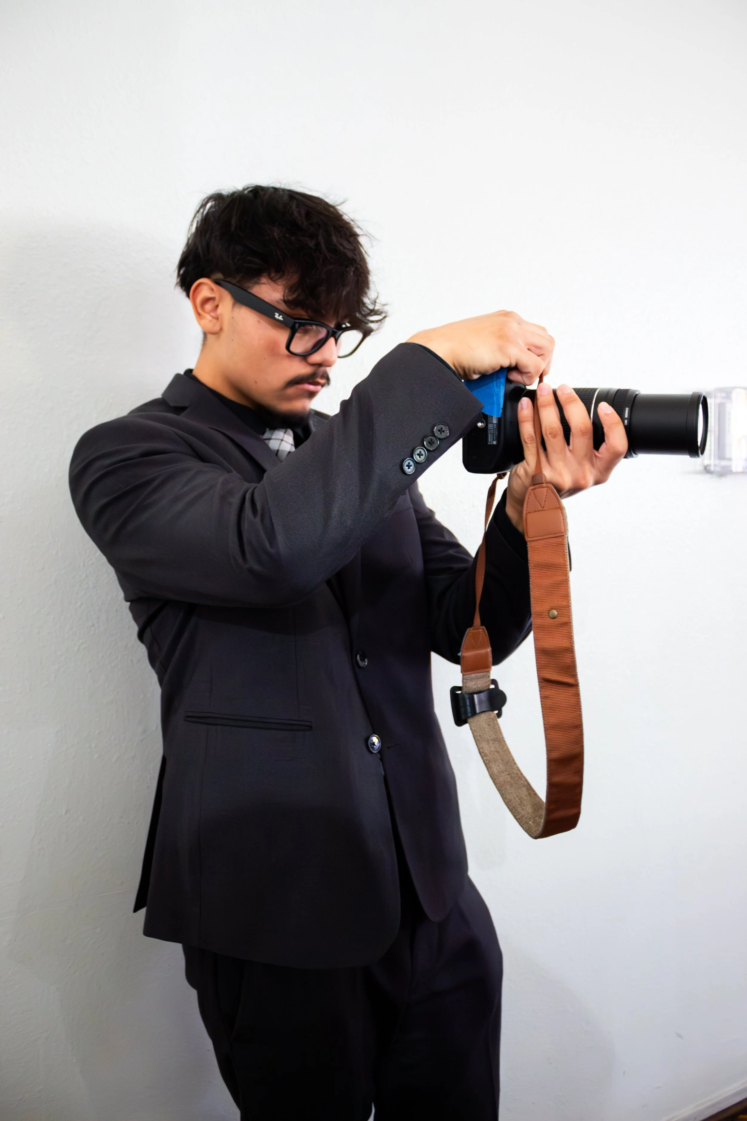 A man in a black suit and glasses is aiming a camera with a long lens at a white wall.