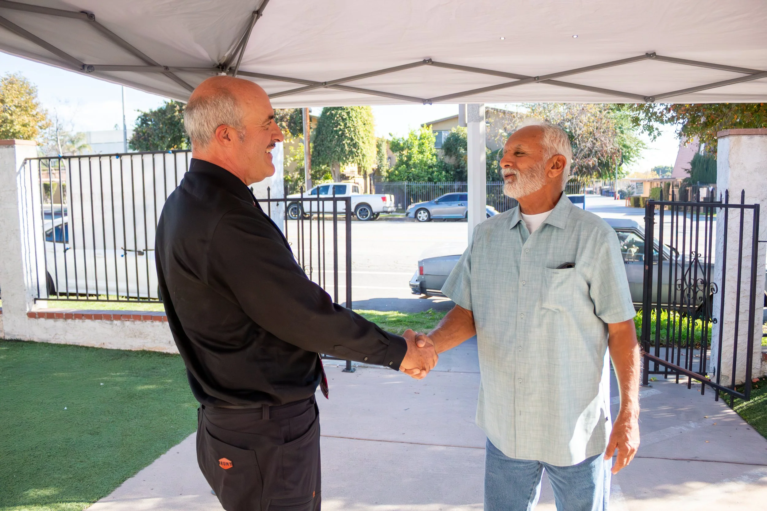 Two men shaking hands outside under a canopy, with a street and parked cars in the background.