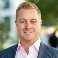 Headshot of a young man with blonde hair, wearing a navy blazer and pink shirt, smiling outdoors.