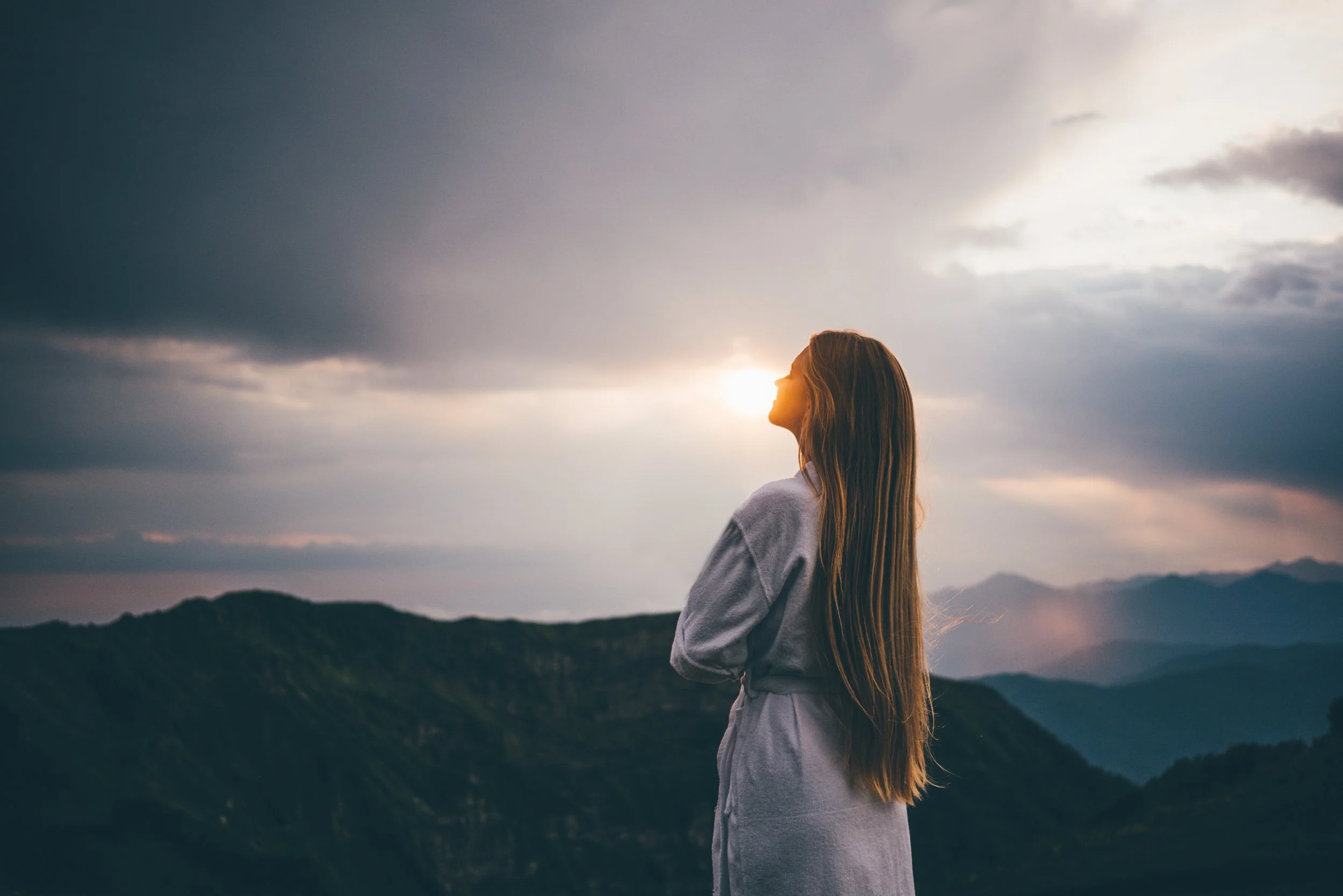 A woman with long hair in a light grey coat stands on a mountain overlooking a landscape of rolling hills,