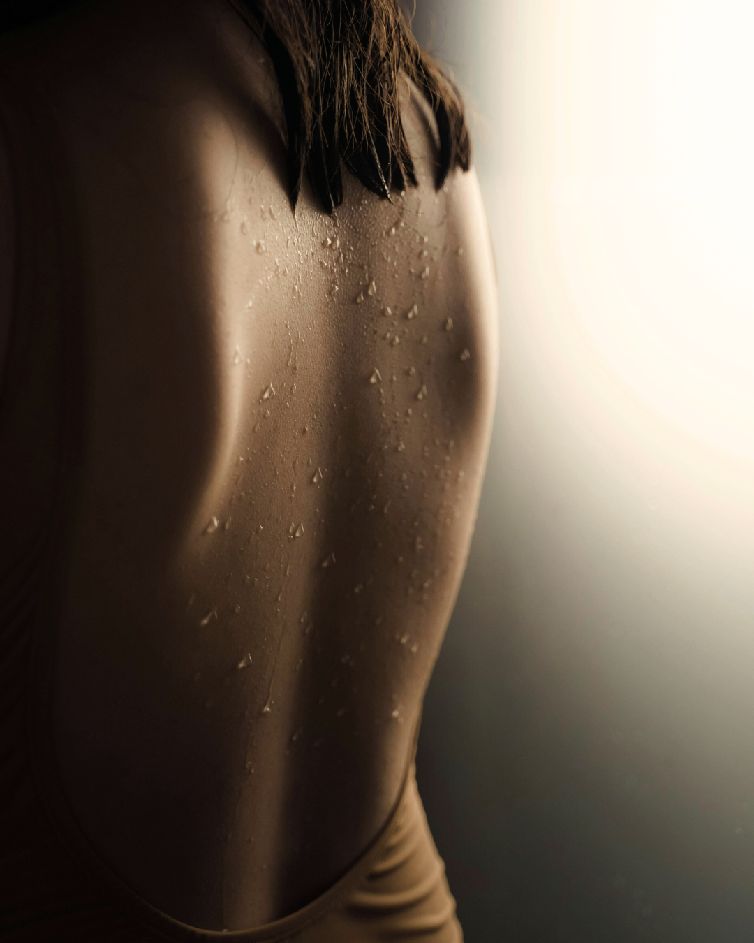 Close-up of a person's shoulder and back with water droplets on skin, with soft lighting highlighting contours and wetness.
