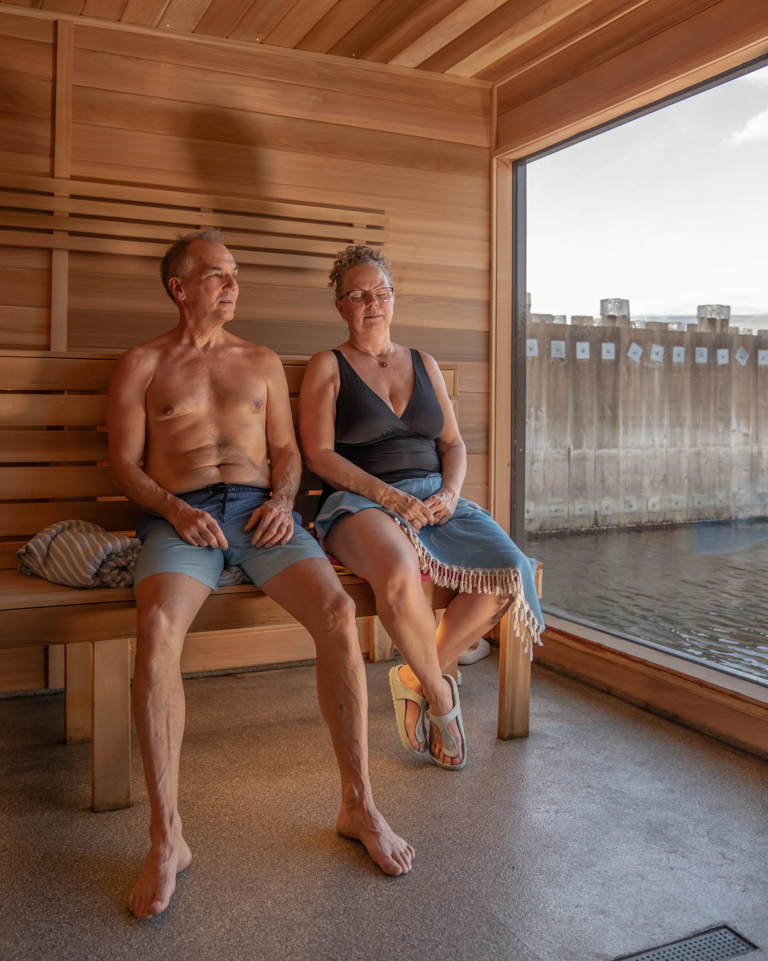 A man and woman sitting inside a wooden sauna, looking out the large window towards the water and sky outside.