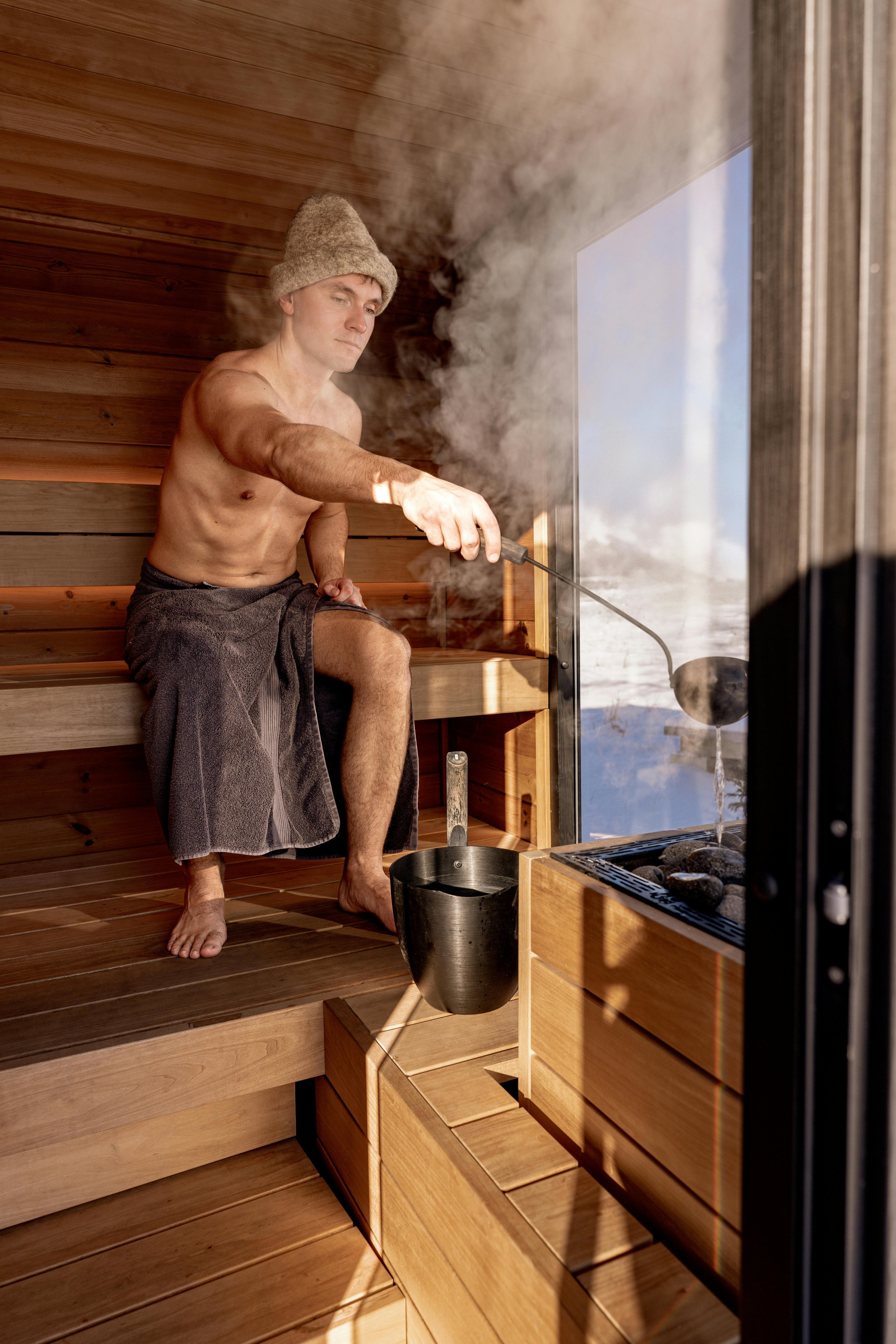 Man in a sauna wearing a towel and a knit cap, using a ladle to pour water on hot stones inside a wooden sauna.