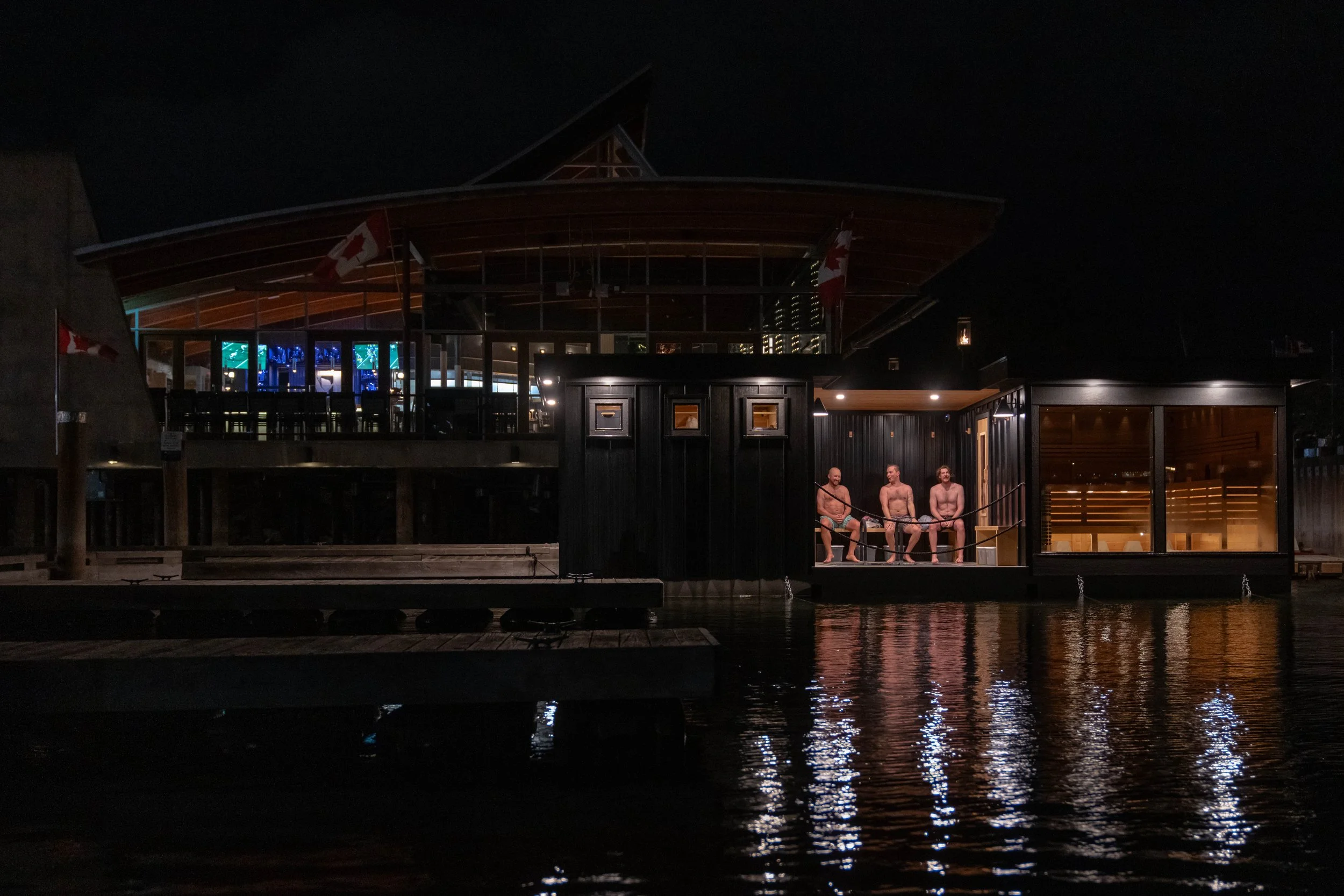 Nighttime scene of Shelter Floating Sauna, with three men relaxing on a dock.