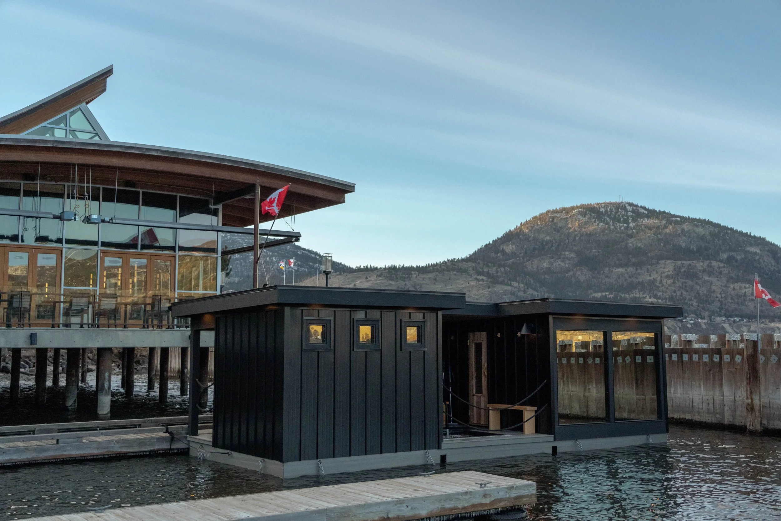 A waterfront building in Penticton, BC, with large glass windows, a Canadian flag, and a mountainous landscape in the background.
