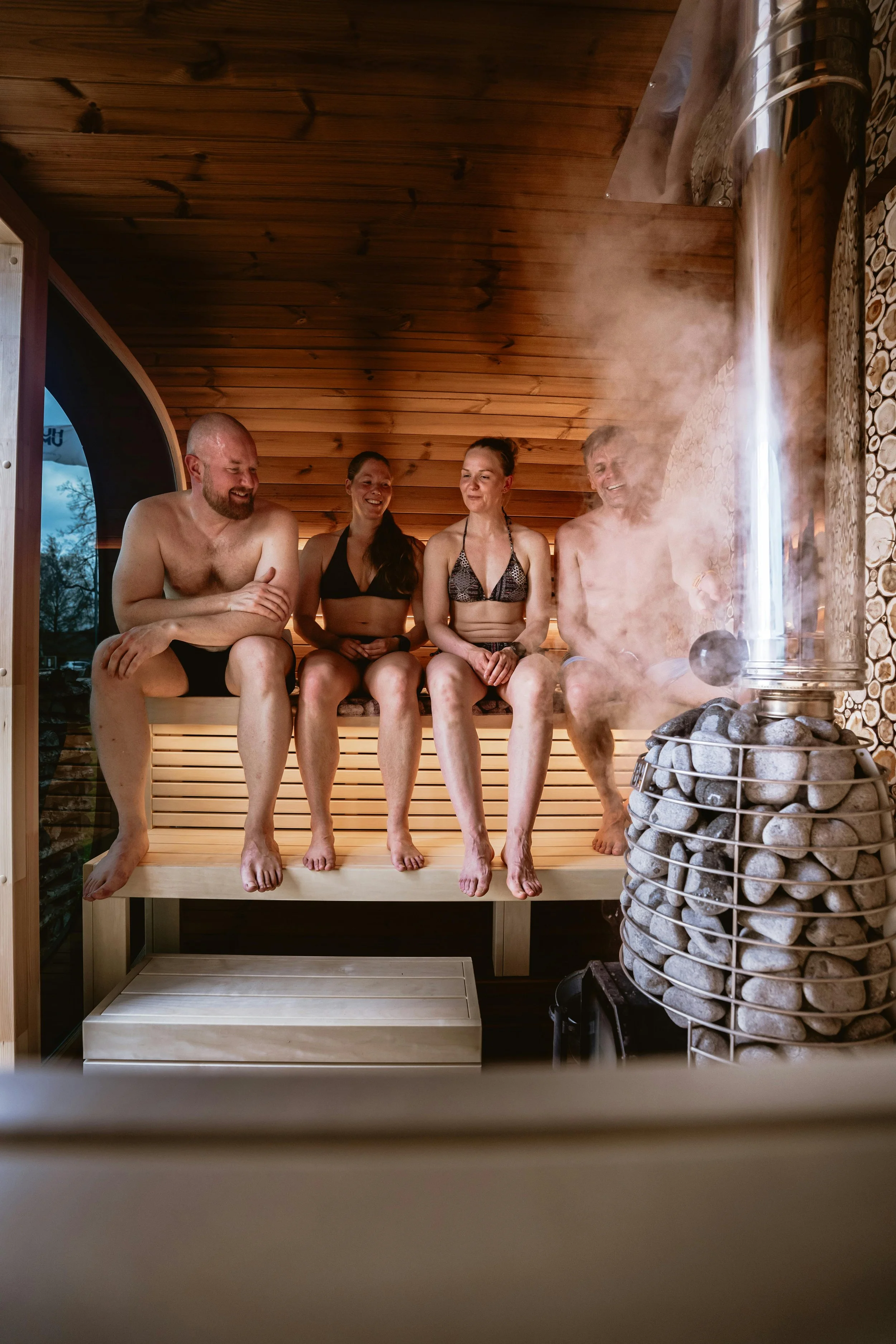 Four people in swimwear sitting on a wooden bench inside a sauna, with one person smiling and looking at the others, steam rising from the sauna heater.