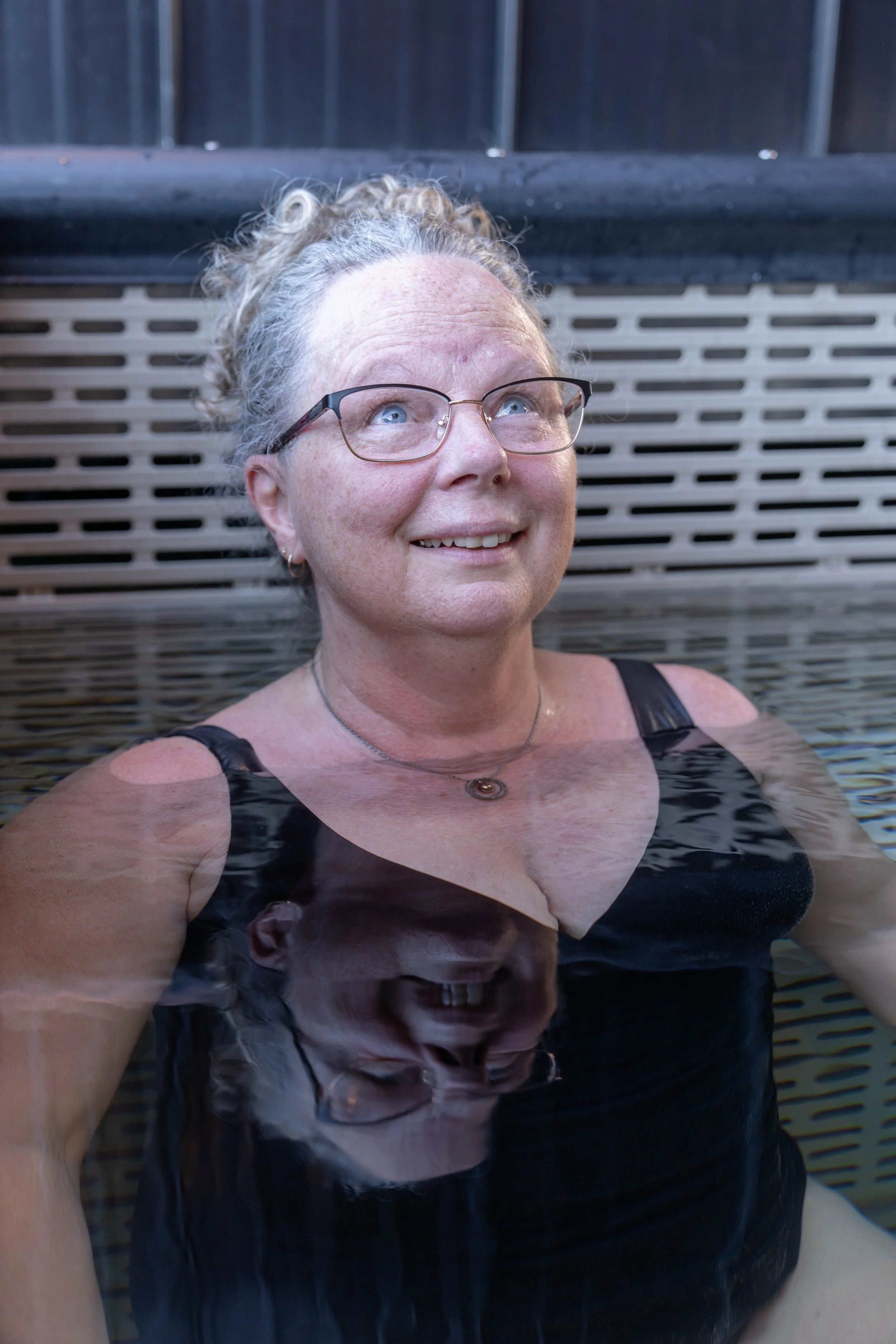 A woman with glasses, relaxing in a cold plunge in Okanagan Lake, looking upward with a smile.
