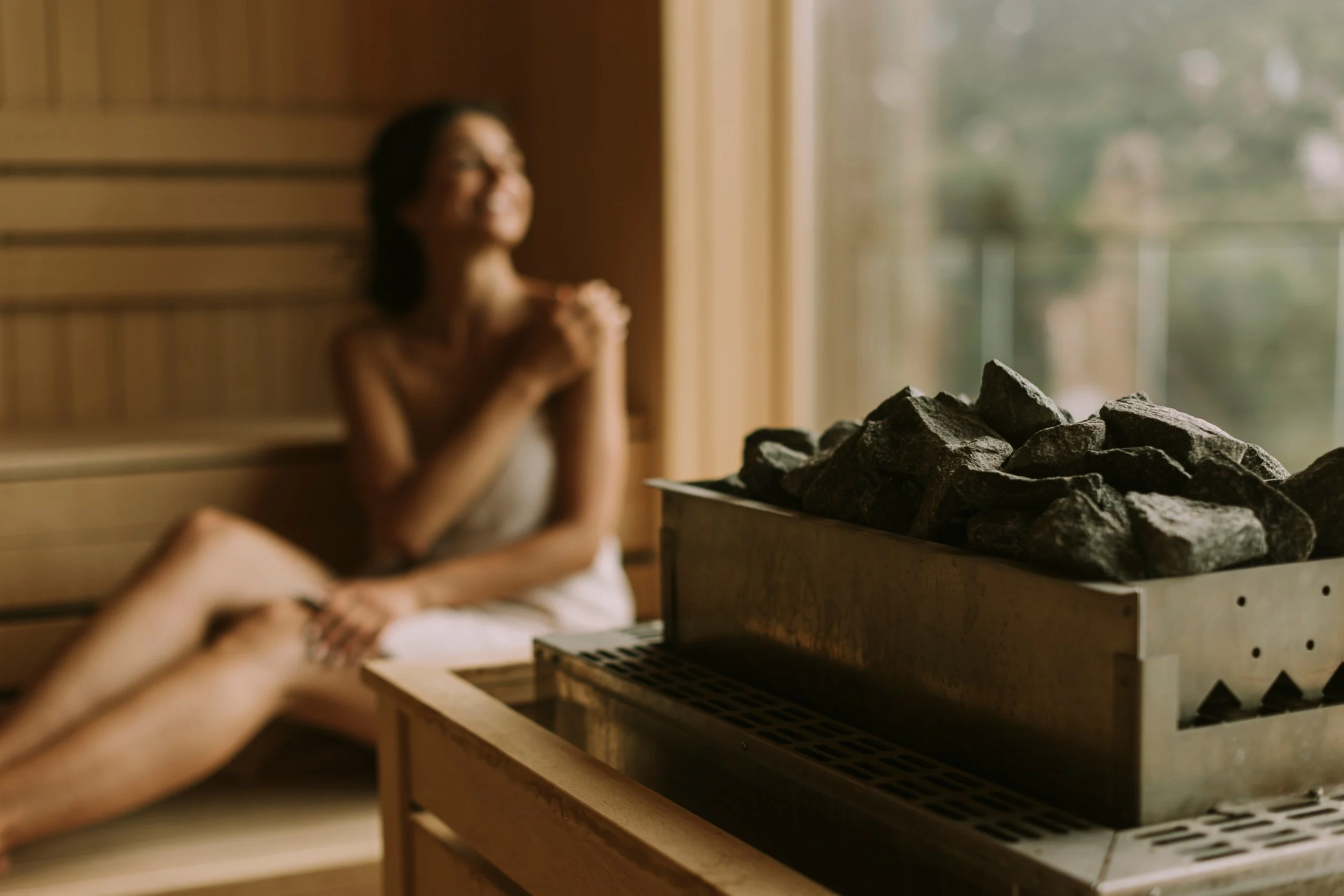 A woman sitting in a sauna, wearing a towel, with a sauna heater filled with rocks visible in the foreground.