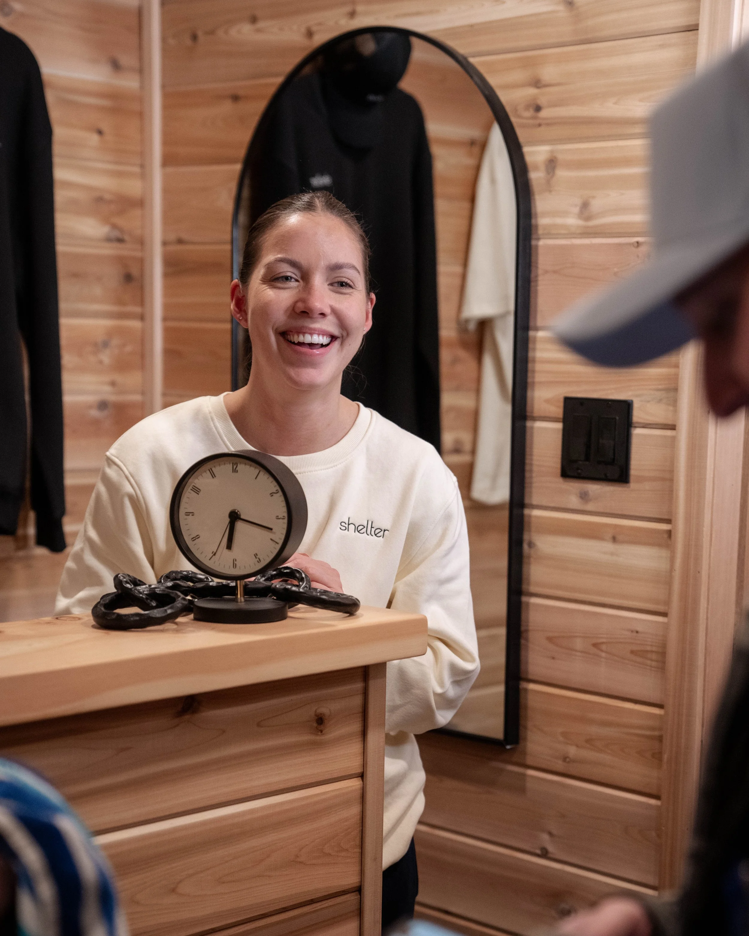A woman with a big smile behind a wooden reception desk, wearing a cream-colored sweatshirt with 'shelter' written on it.