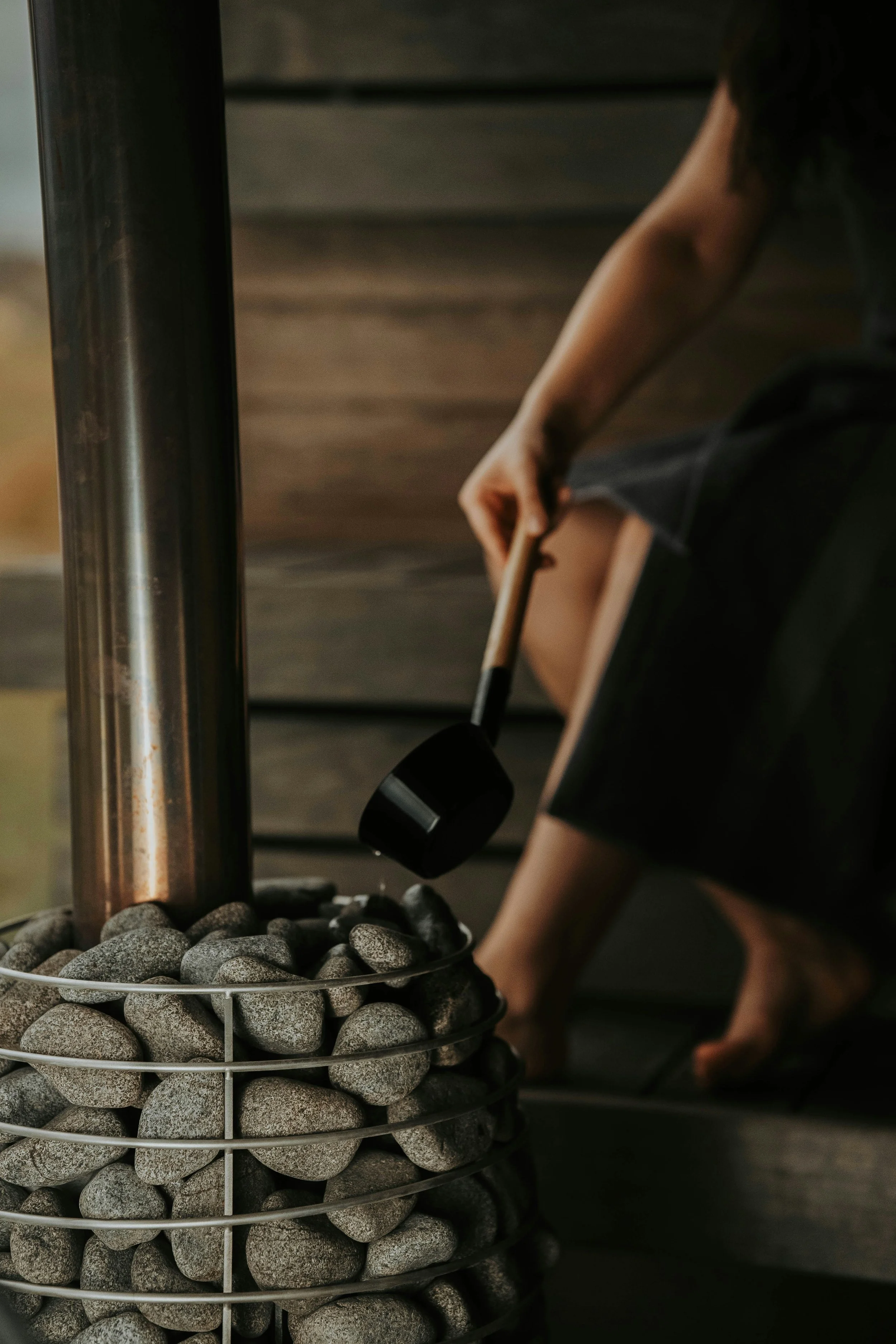 Person sitting outdoors, stirring a container of smooth gray stones with a black ladle.
