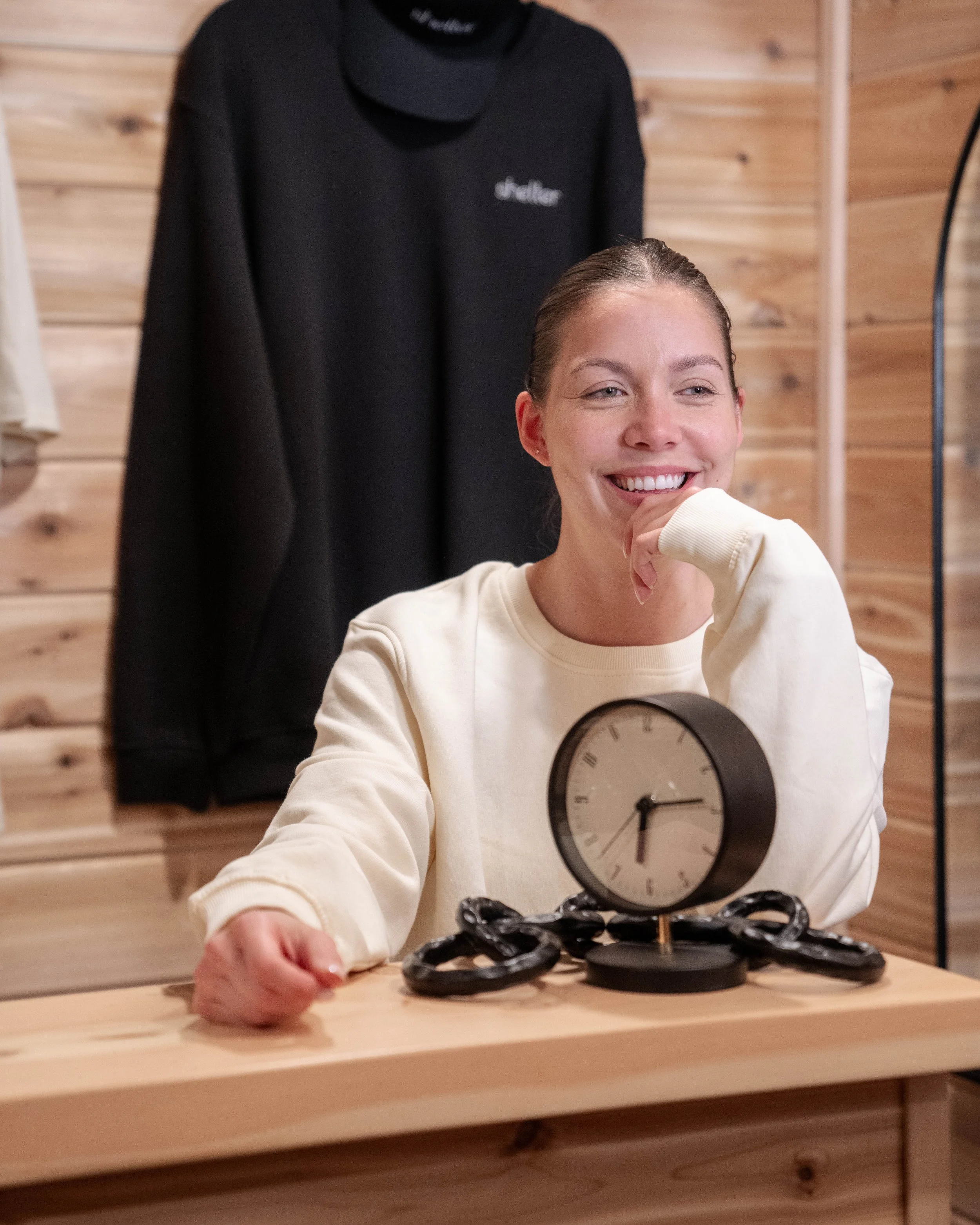 A woman smiling and sitting at a wooden table with a black clock and black chains, with a black hoodie hanging on the wooden wall behind her.