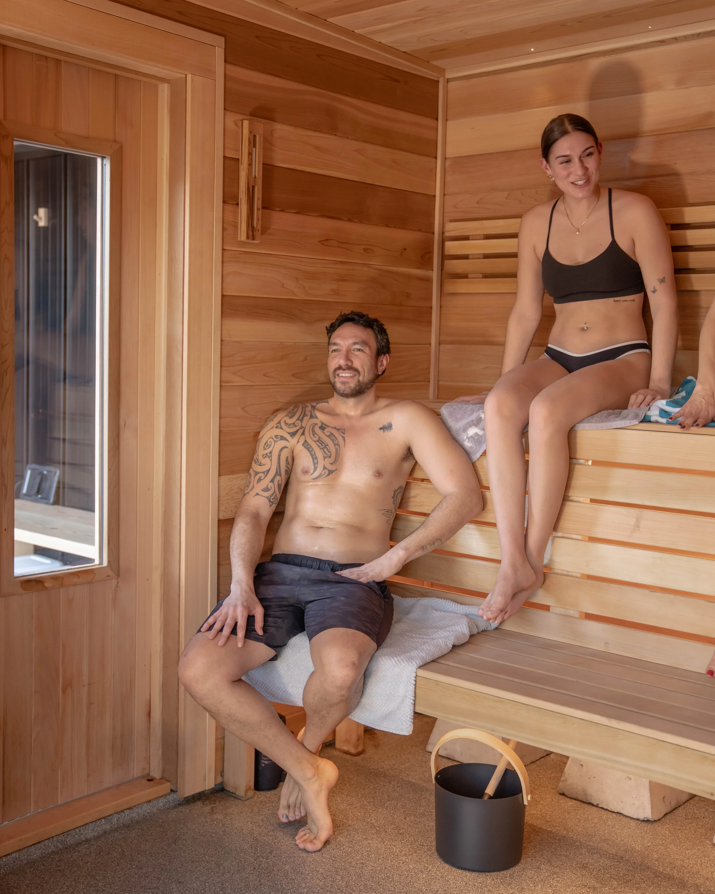 A man and woman are relaxing inside a wooden sauna; at Shelter Floating Sauna in Penticton, BC.
