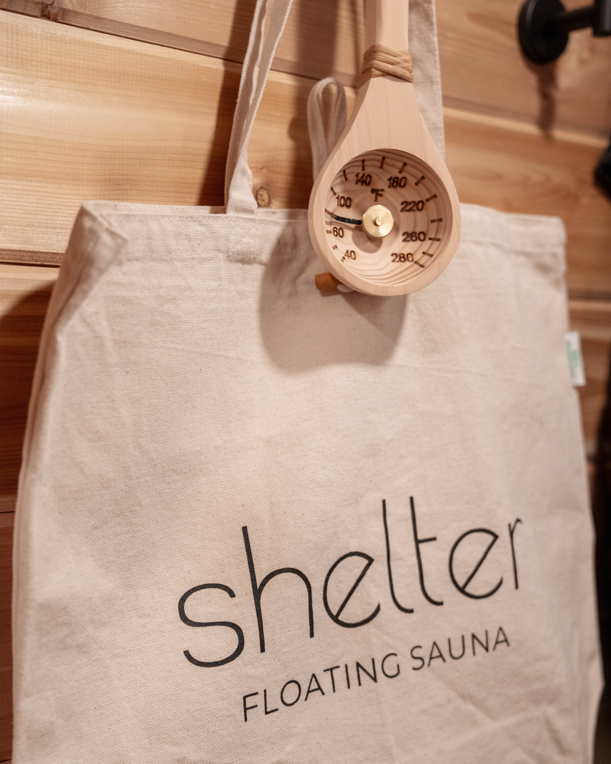 A white tote bag with 'Shelter Floating Sauna' printed on it, hung on a wooden wall.