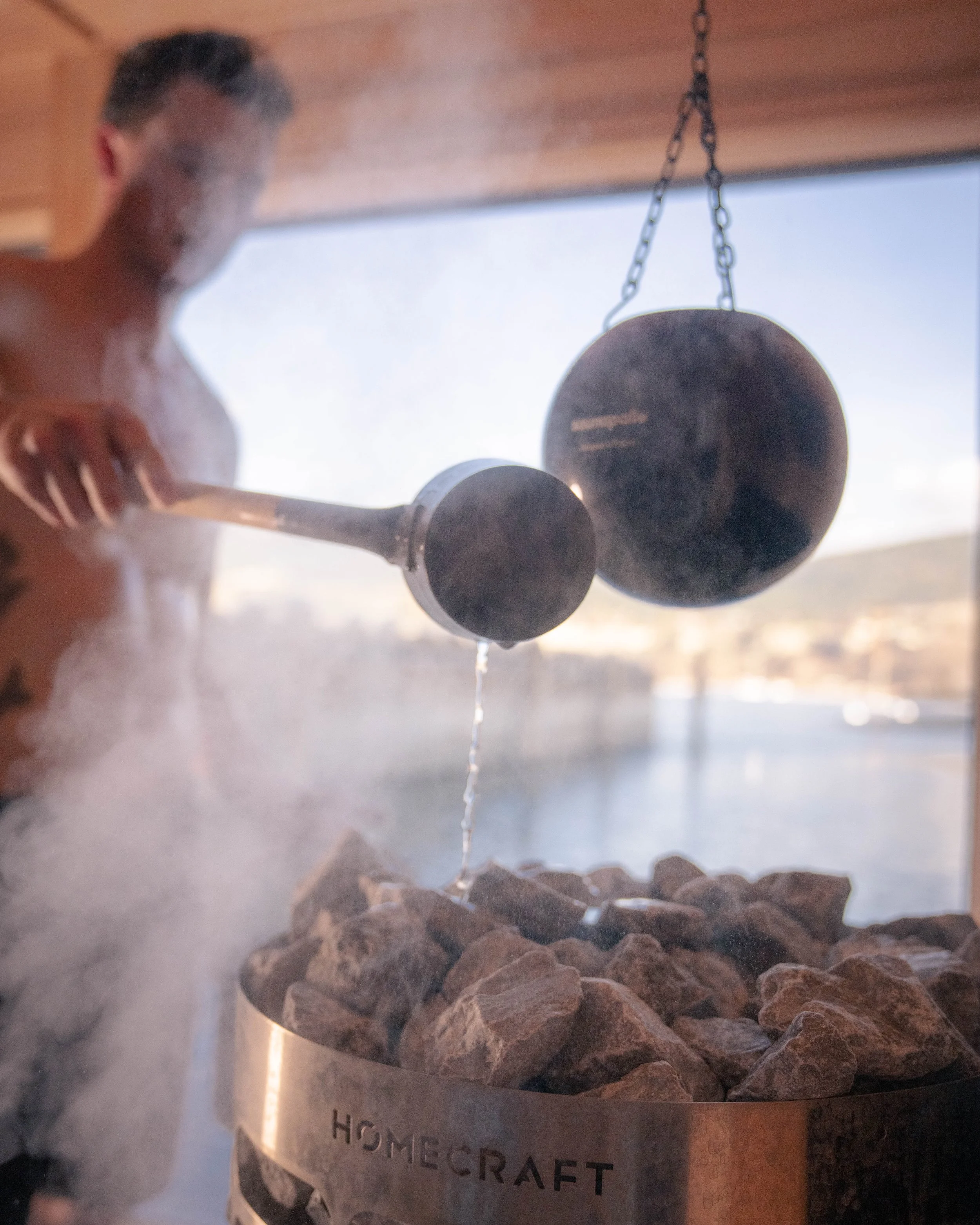 A person is pouring water into a rock-filled container at a sauna or spa, with steam rising.