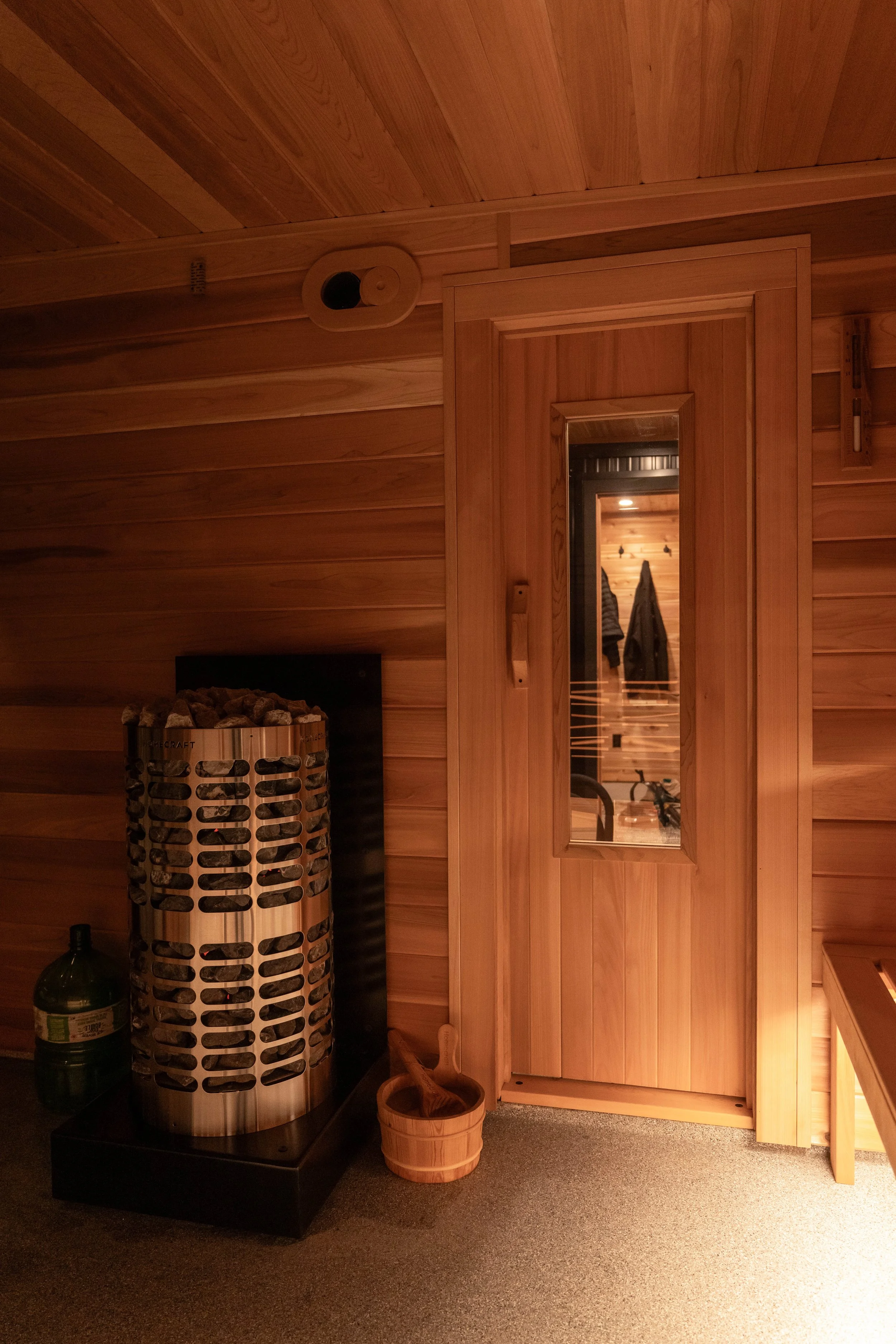 Wood-paneled sauna room with a door, a wooden bucket and ladle, a bottle of water, and a sauna heater.