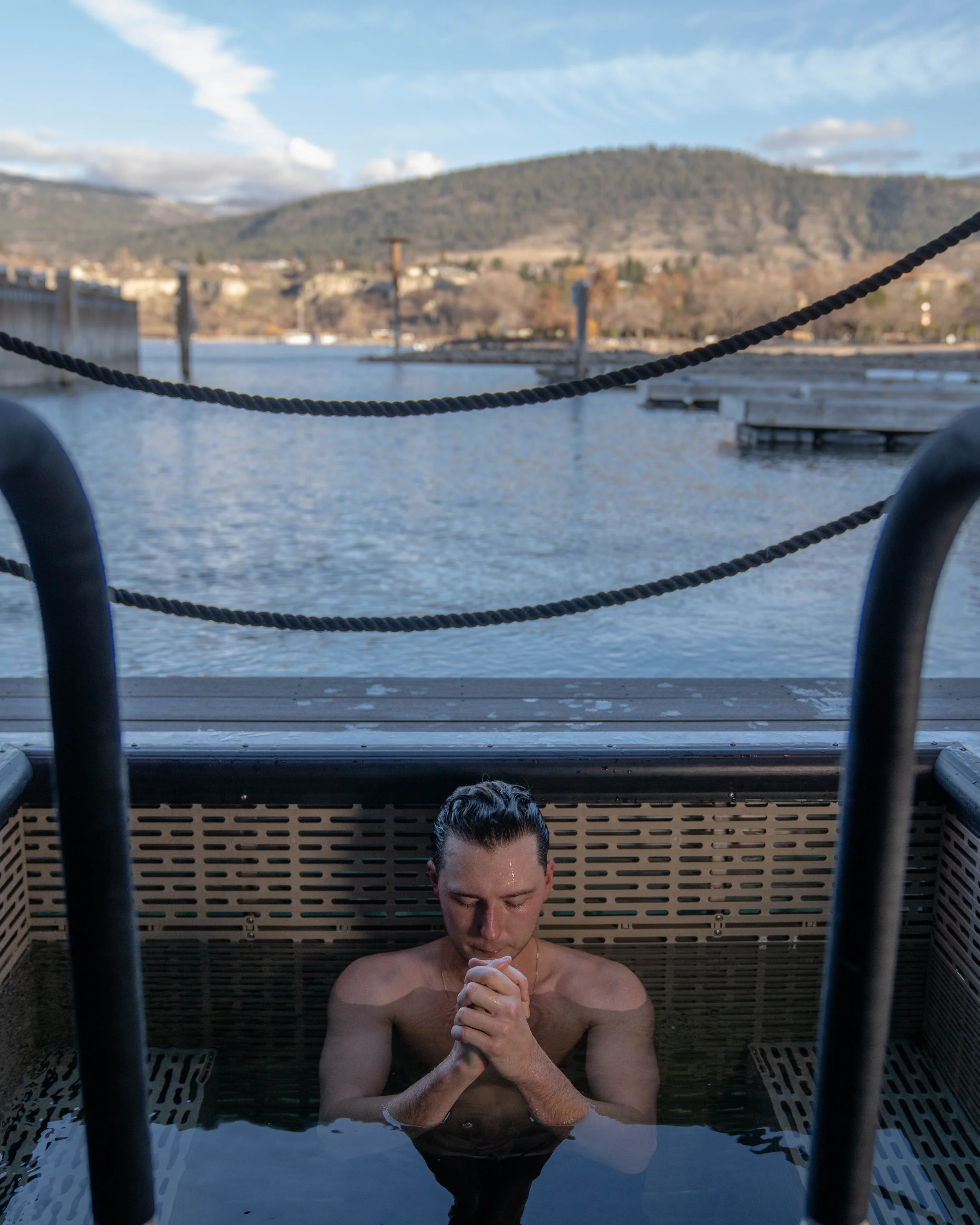 A man with wet hair in an outdoor cold plunge near a body of water, with Okanagan Lake and cloudy sky in the background.