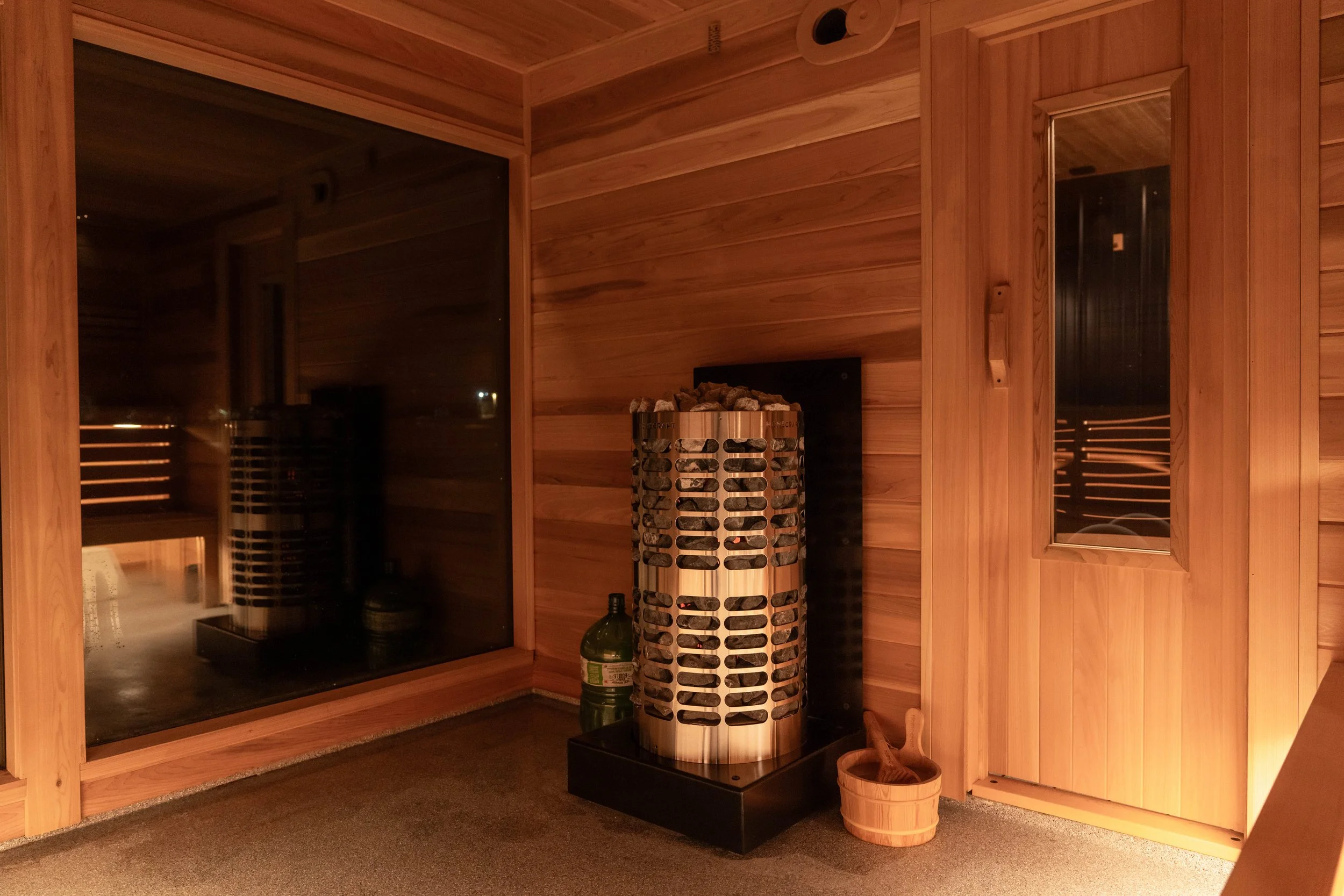 Indoor sauna room with wooden walls and door, large window, heater with stones, and a small bucket with a ladle.