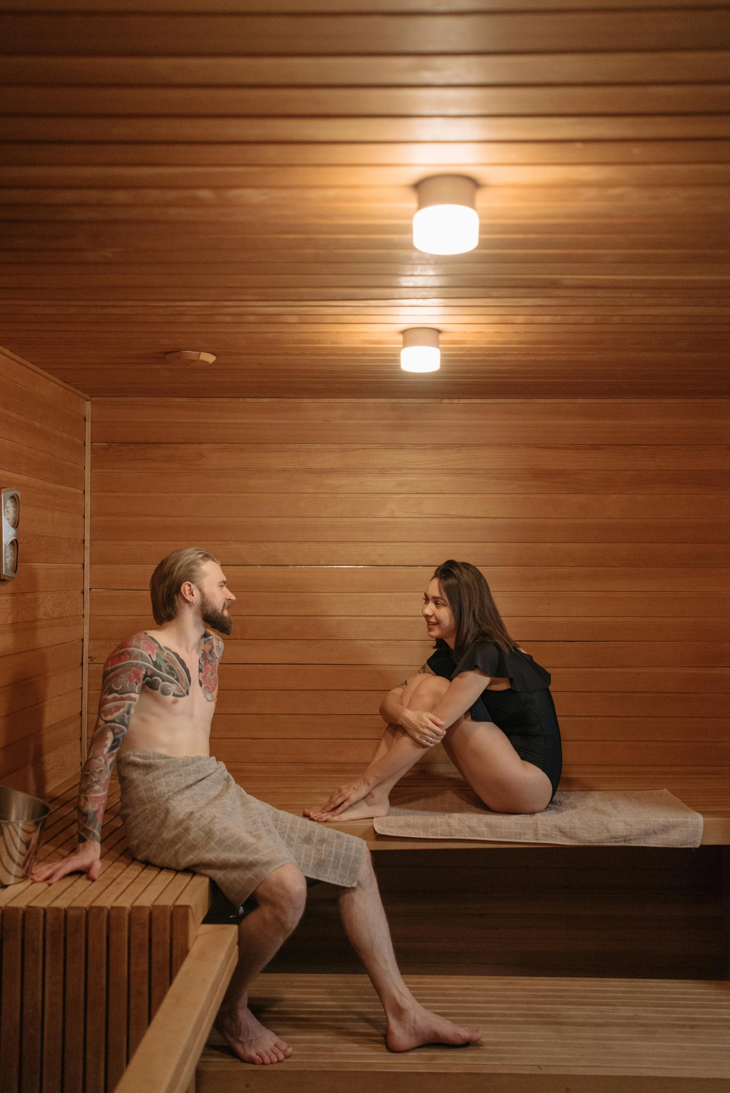 A man and woman sitting in a wooden sauna, engaging in conversation and smiling at each other.